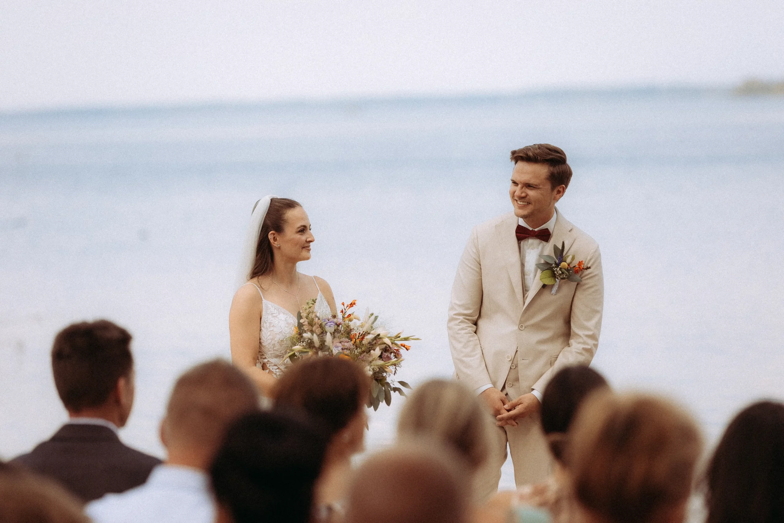 Brautpaar am Strand, die Braut mit Blumenstrauß blickt liebevoll zum Bräutigam. Hochzeitsfotograf in MV hat diese romantische Szene eingefangen. Gäste im Vordergrund.