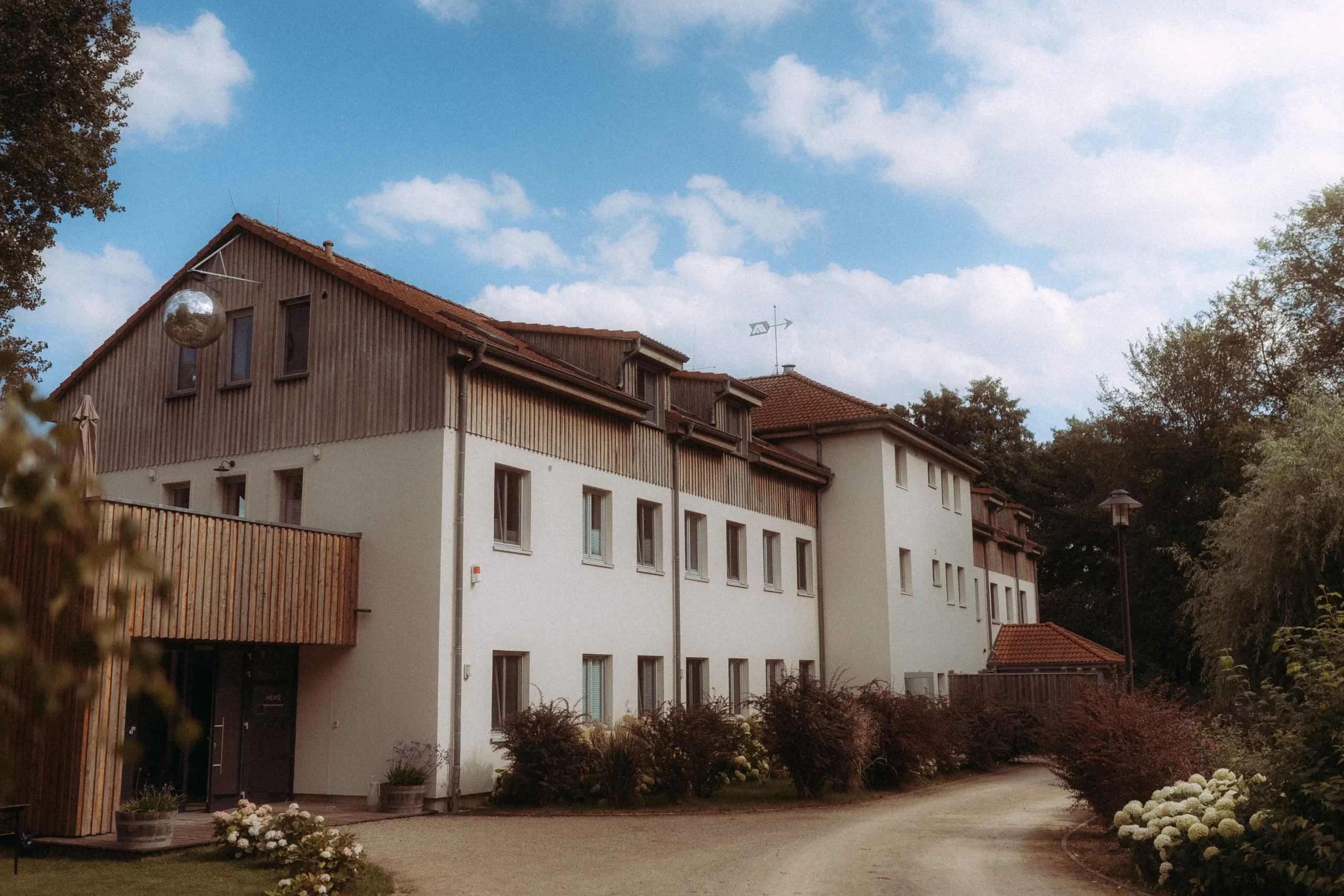 Ein großes, helles Gebäude mit Holzverkleidung unter blauem Himmel. Der Weg führt zum Haus, umgeben von üppigem Grün. Fotograf in MV könnte hier wundervolle Familienfotos MV machen.