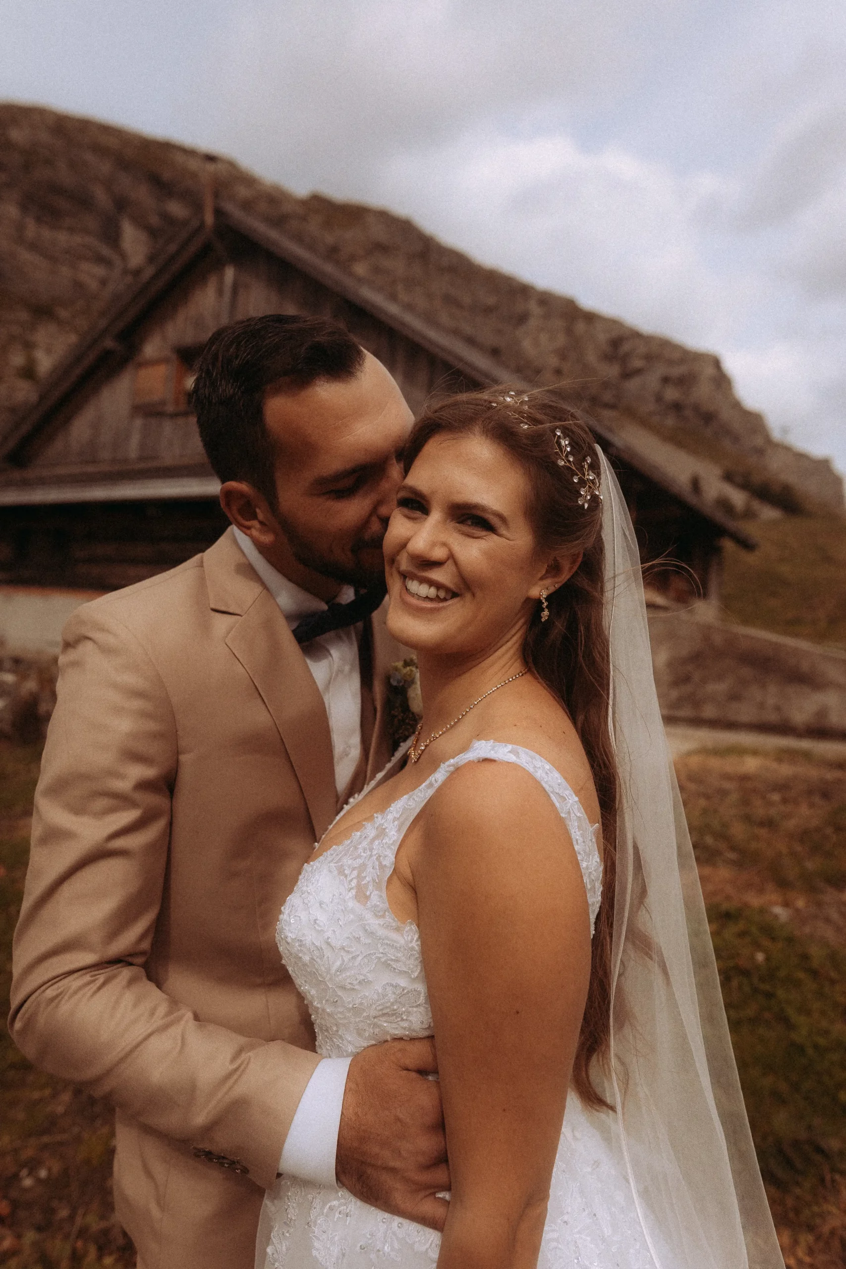 Ein strahlendes Brautpaar, der Bräutigam küsst die Braut zärtlich. Ländliche Hochzeit vor einer Hütte. Hochzeitsfotograf in Schwerin hat diesen besonderen Moment eingefangen.