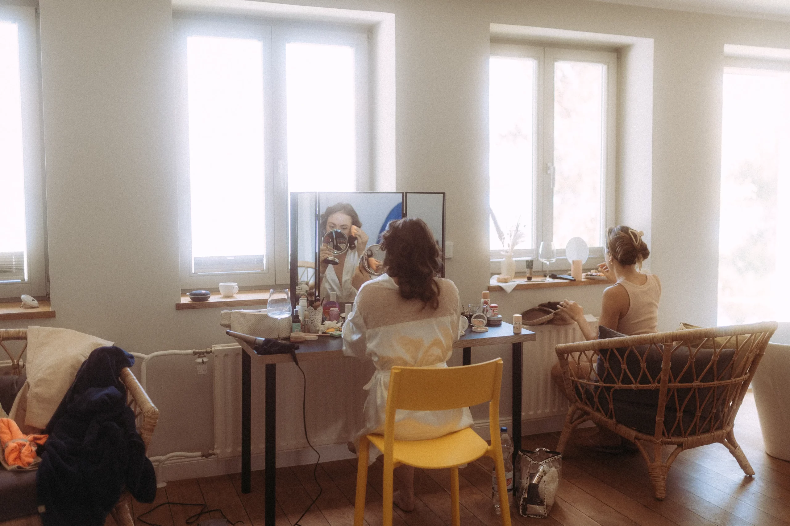 Zwei Frauen sitzen in einem hellen Raum mit großen Fenstern; eine steht vor einem Spiegel und macht ein Selfie, während die andere auf einem geflochtenen Stuhl am Fenster sitzt. Make-up und persönliche Gegenstände liegen auf dem Tisch verstreut - perfekt für einen Hochzeitsfotografen in Schwerin.