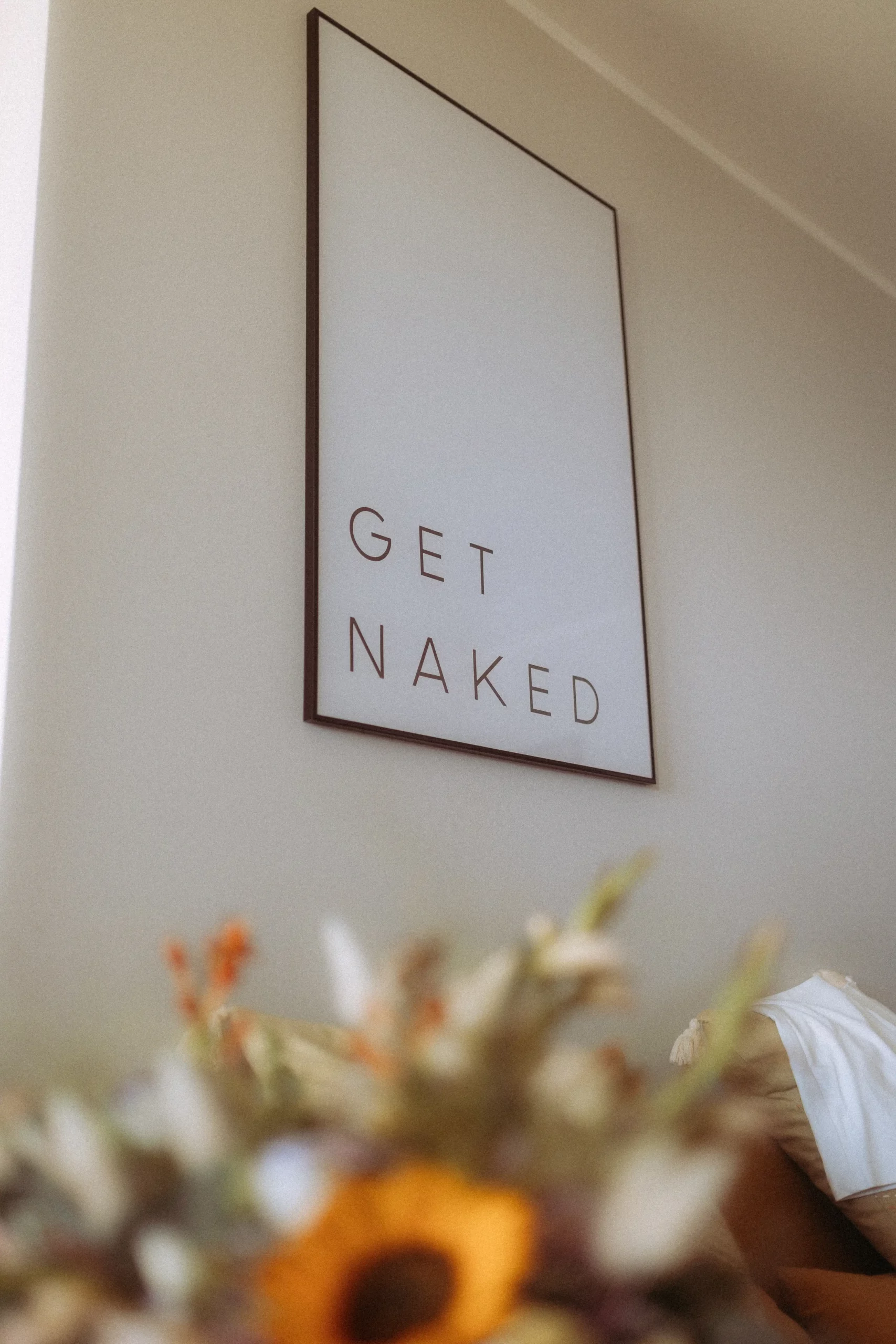 Ein gerahmtes Schild an einer beigen Wand zeigt in großen, schlichten Buchstaben die Worte GET NAKED. Im Vordergrund sind verschwommene Blumen zu sehen, die ein modernes Ambiente schaffen, das perfekt für einen Fotografen in Schwerin ist.