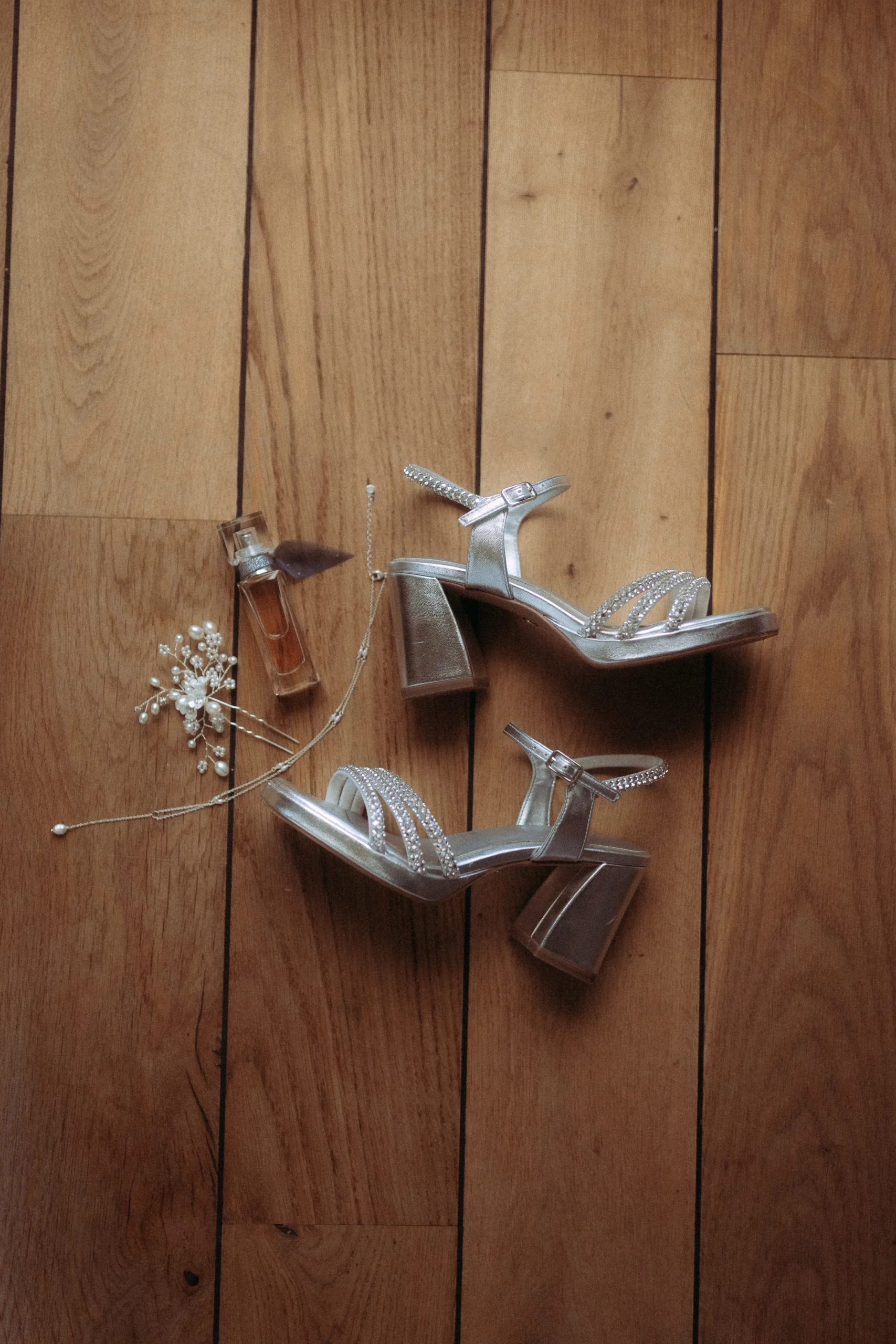 Silberne hochhackige Sandalen, ein kleines Parfümfläschchen und ein zarter Perlenhaarschmuck auf einem Holzboden - perfekte Details, die ein Hochzeitsfotograf in Schwerin eingefangen hat.
