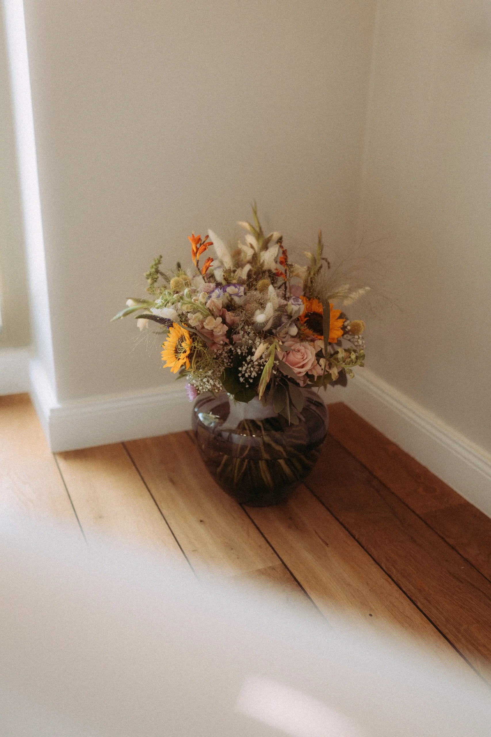 Eine Glasvase, gefüllt mit einem Strauß bunter Blumen, darunter auch Sonnenblumen, steht auf einem Holzboden in der Ecke eines sonnendurchfluteten Raums mit weißen Wänden - wunderschön eingefangen von einem Fotografen in Schwerin.