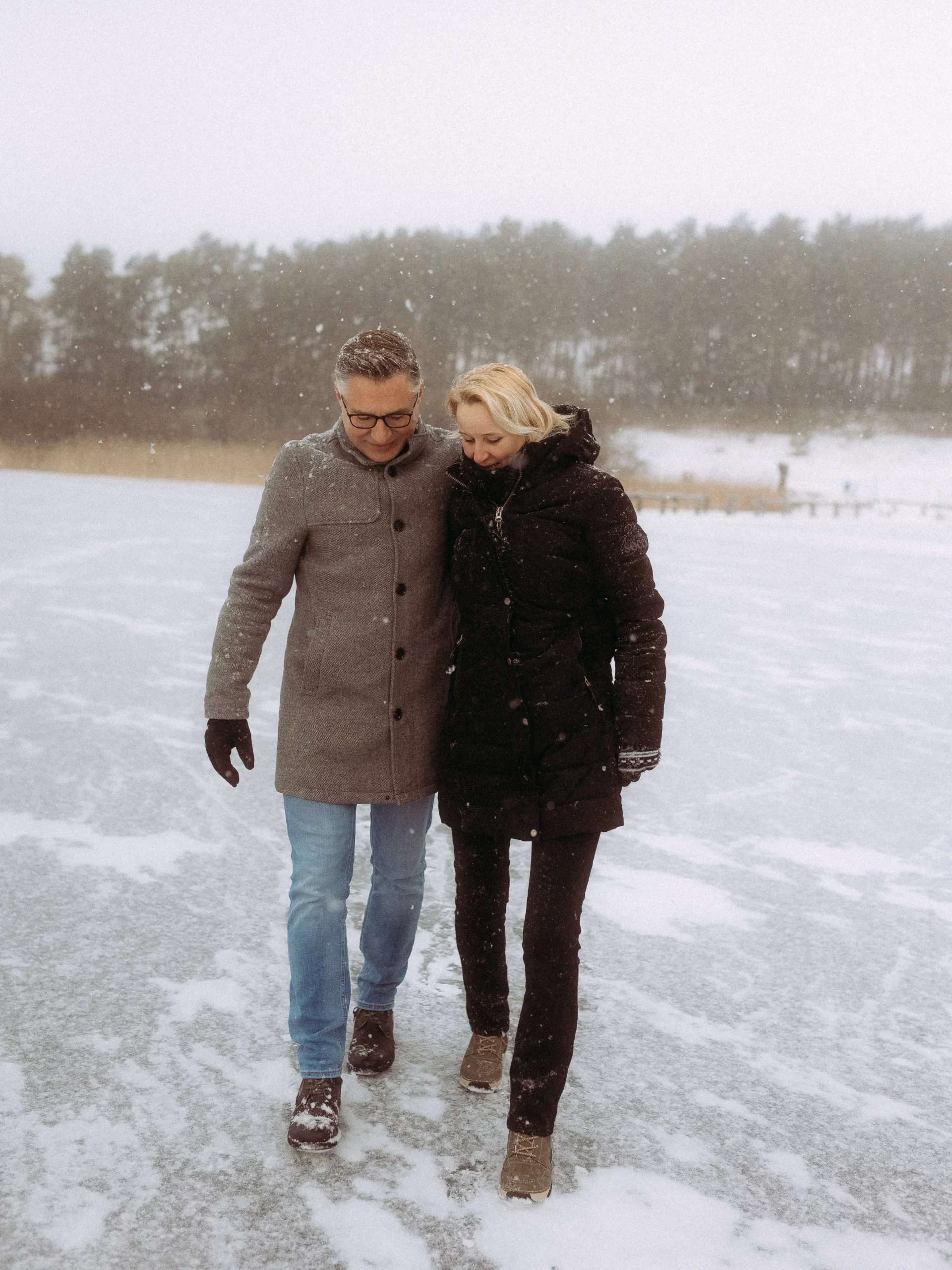 Ein Mann und eine Frau gehen eng beieinander durch eine verschneite Landschaft, warm gekleidet in Wintermäntel und Stiefel, mit Bäumen und Schneefall im Hintergrund - perfekt für unvergessliche Hochzeitsfotos MV.