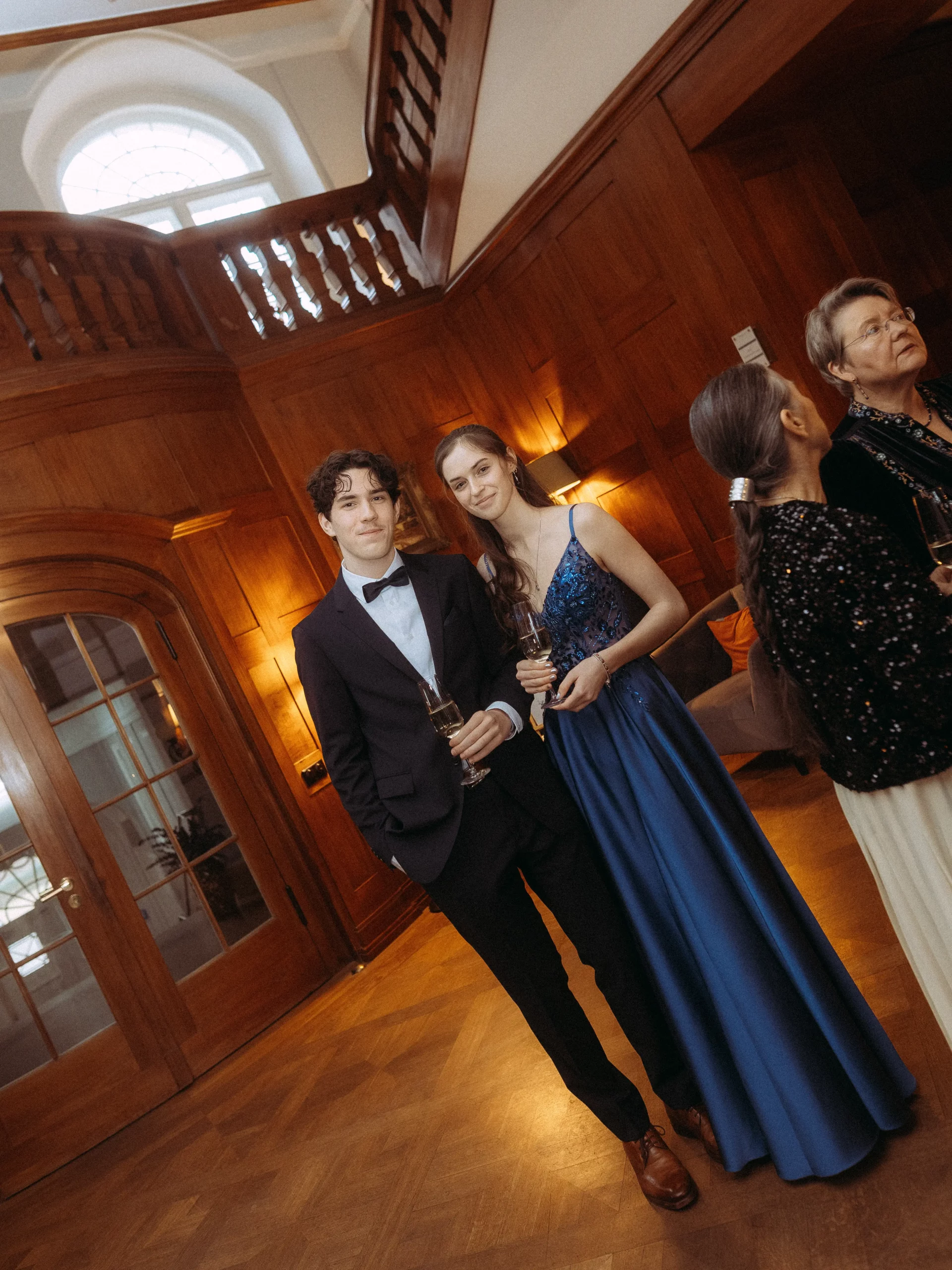 Ein junger Mann im Smoking und eine junge Frau in einem blauen Abendkleid stehen bei einer eleganten Veranstaltung in einem holzgetäfelten Raum zusammen und halten Gläser in der Hand. In der Nähe unterhalten sich die Gäste angeregt. Hochzeitsfotograf aus Schwerin hält die besonderen Momente fest.