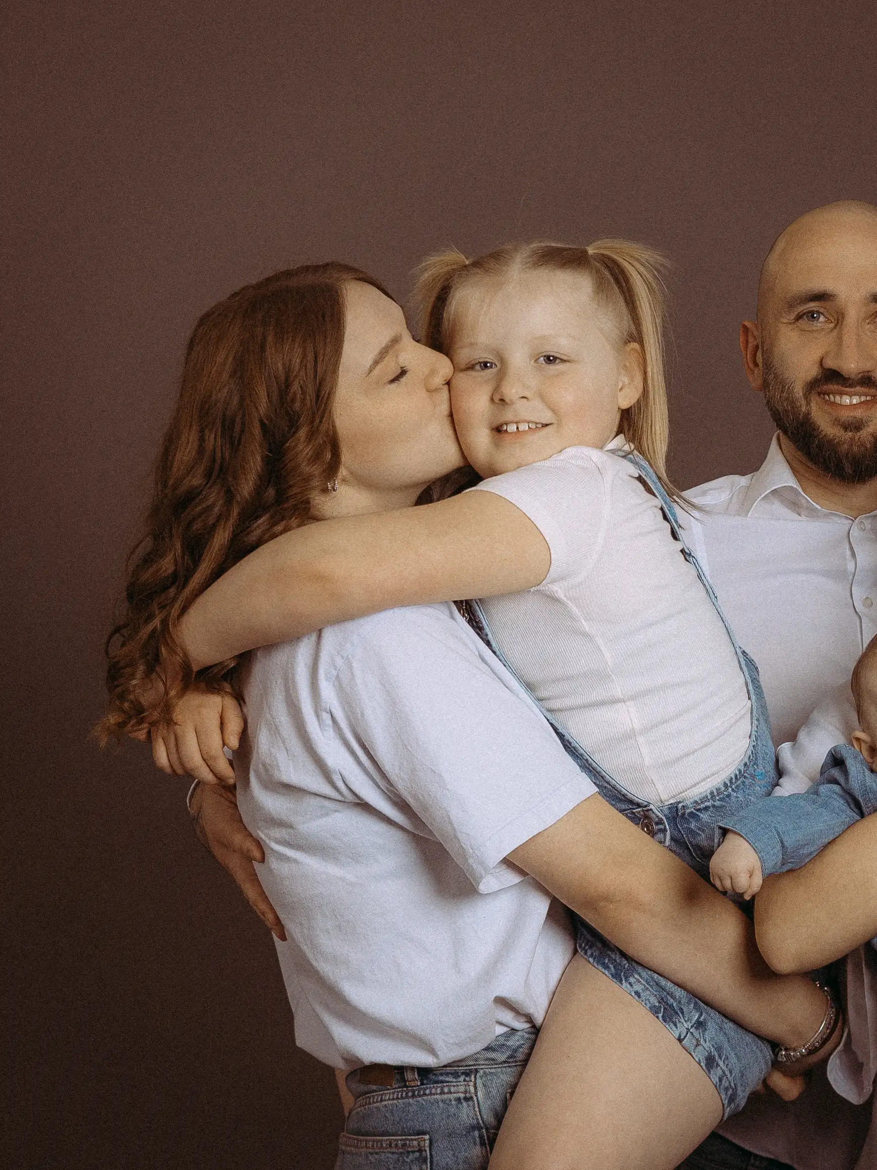 Familienfoto MV: Mutter umarmt lächelnde Tochter, gibt Kuss. Vater und Baby im Hintergrund. Liebevolle Familienszene, aufgenommen vom Fotograf aus Schwerin.