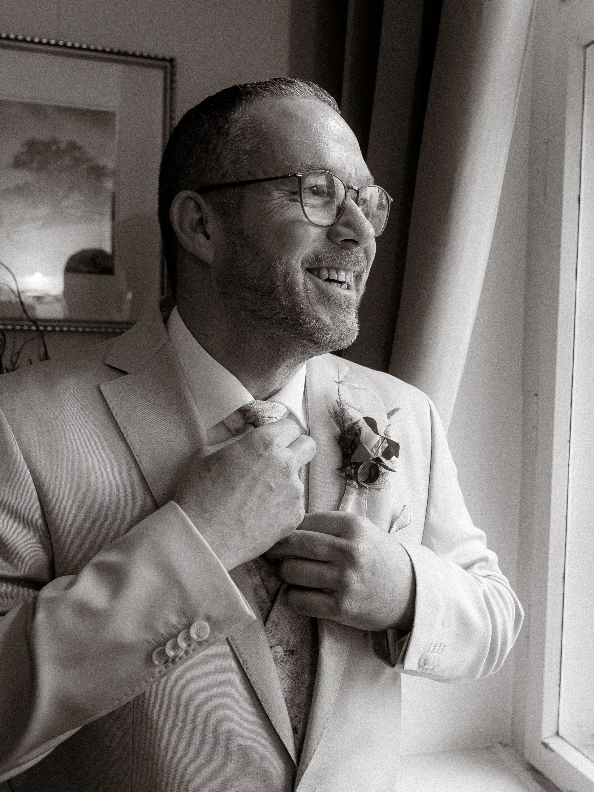 Ein Mann in einem hellen Anzug und mit Brille lächelt, während er seine Krawatte zurechtrückt. Er steht an einem Fenster mit natürlichem Licht. Aufgenommen von Hochzeitsfotograf MV, trägt er eine Boutonniere am Revers. Hinter ihm hängt ein gerahmtes Bild an der Wand in diesem Schwarz-Weiß-Bild.