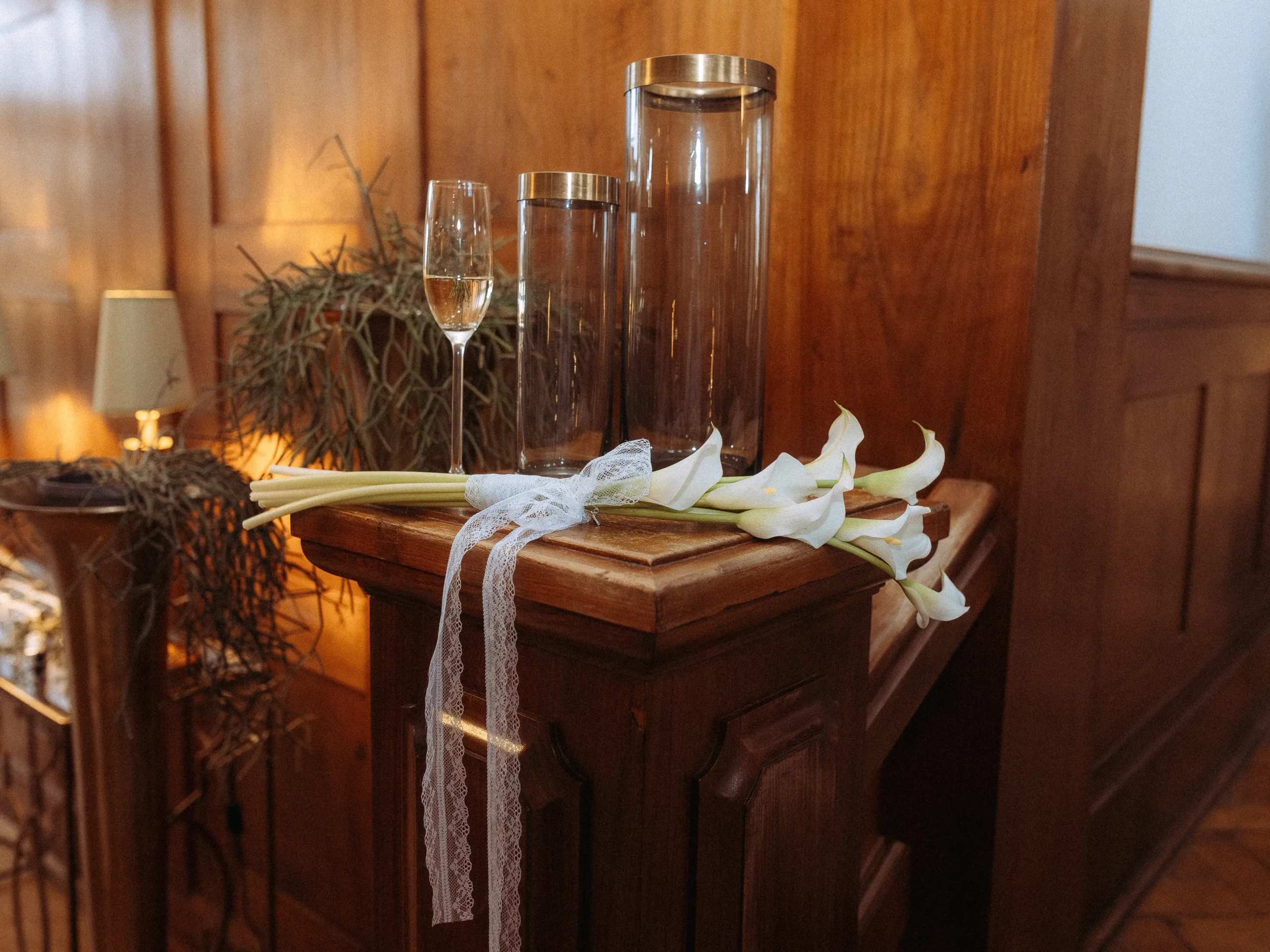 Ein kleiner Strauß weißer Calla-Lilien, mit Spitze gebunden, liegt auf einem Holzsims neben drei Glasbehältern und einer Sektflöte, mit Holzvertäfelung und sanfter Beleuchtung - die perfekte Inspiration für Hochzeitsfotos MV.