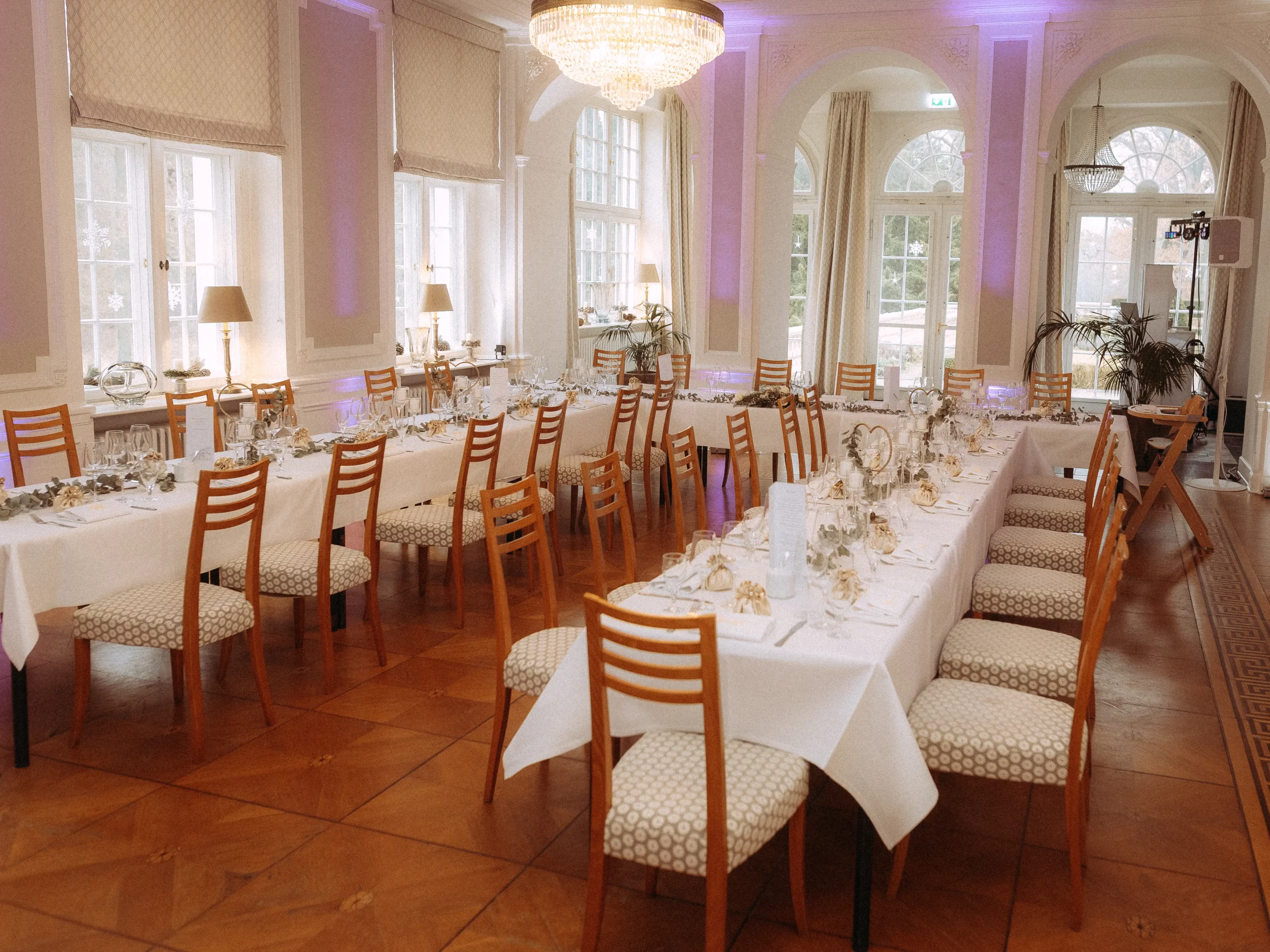 Elegantes Esszimmer für eine Veranstaltung mit weißen Tischdecken, Holzstühlen, Glaswaren und Tischdekoration. Große Fenster, Kronleuchter und sanfte Beleuchtung schaffen eine elegante Atmosphäre - perfekt für Hochzeitsfotos MV.