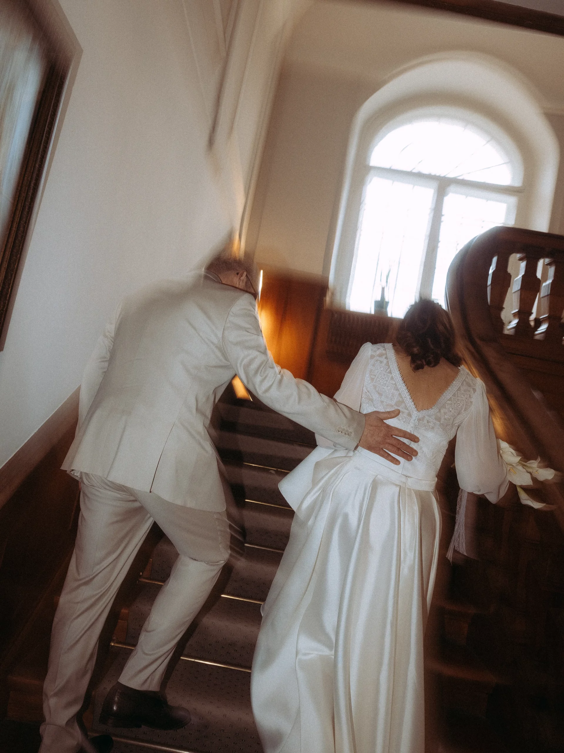 Ein Paar in Hochzeitskleidung, der Bräutigam in einem hellen Anzug und die Braut in einem weißen Kleid, geht eine Holztreppe hinauf zu einem hellen Fenster. Dieses leicht verwackelte Bild fängt authentische Hochzeitsfotos MV von einem Fotografen in MV ein.