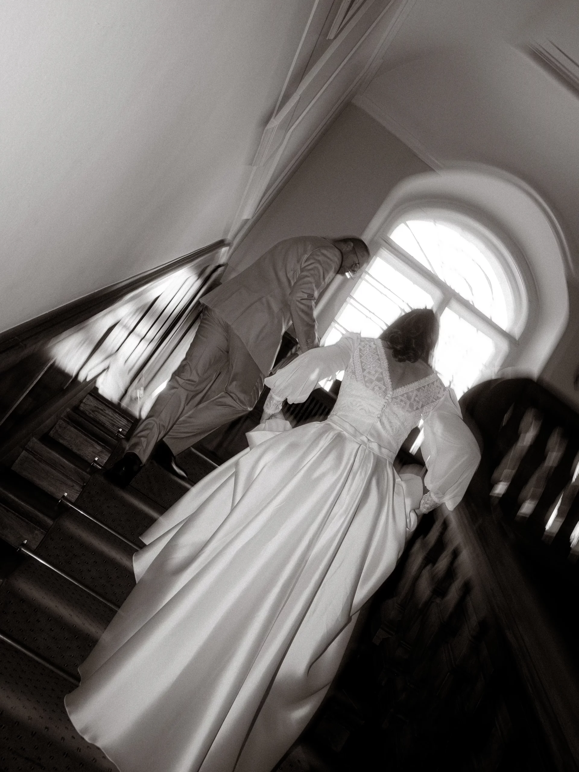Eine Braut in einem langen weißen Kleid und ein Mann im Anzug gehen gemeinsam eine Treppe in einem historischen Gebäude hinauf, von hinten gesehen in schwarz-weiß. Moment eingefangen von Hochzeitsfotograf in MV.