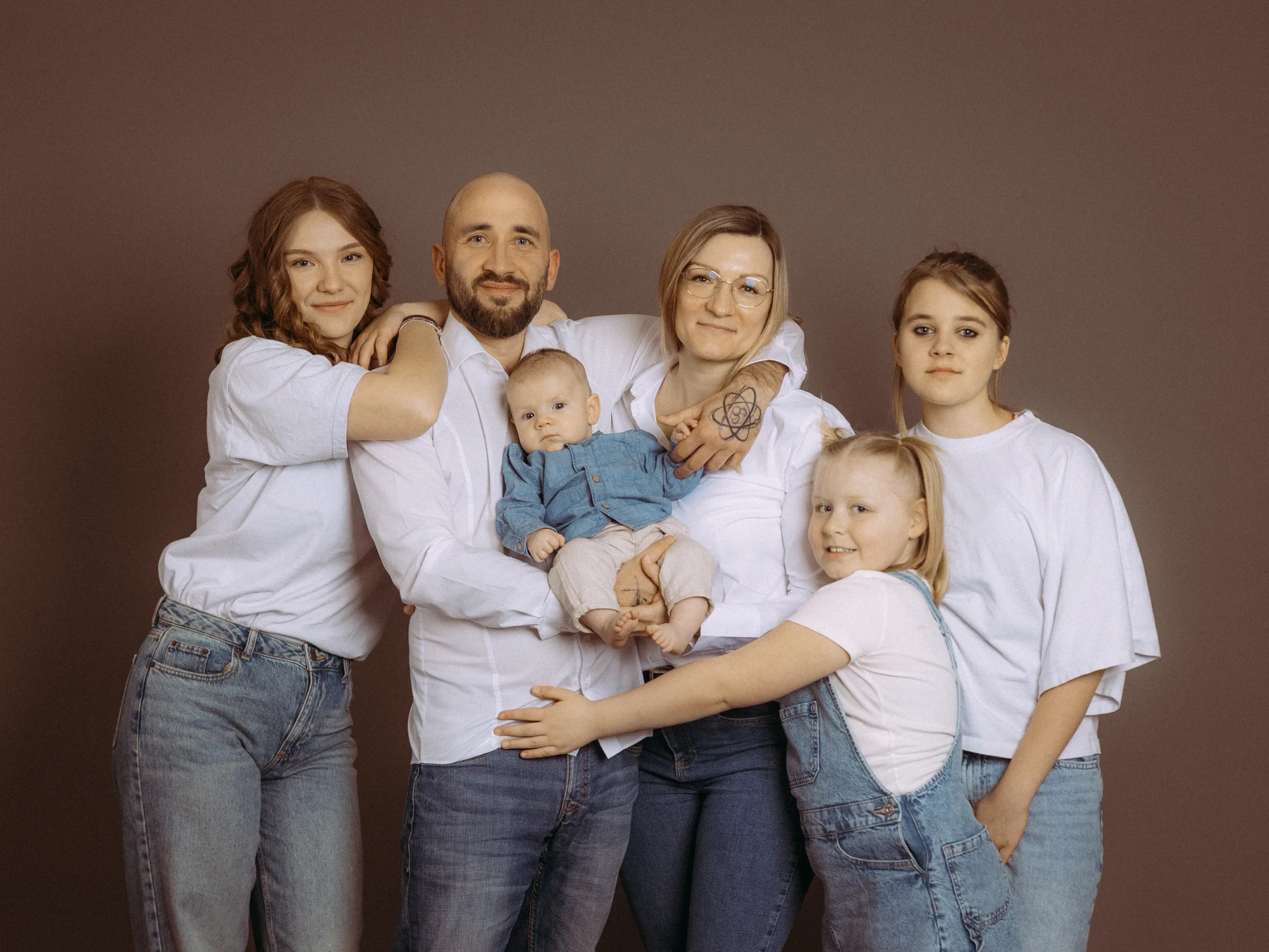 Familienfoto MV: Eine Familie mit fünf Personen, inklusive Baby, posiert in Jeans und weißen Oberteilen vor neutralem Hintergrund. Der Familienfotograf in Schwerin hat die Gruppe liebevoll arrangiert.