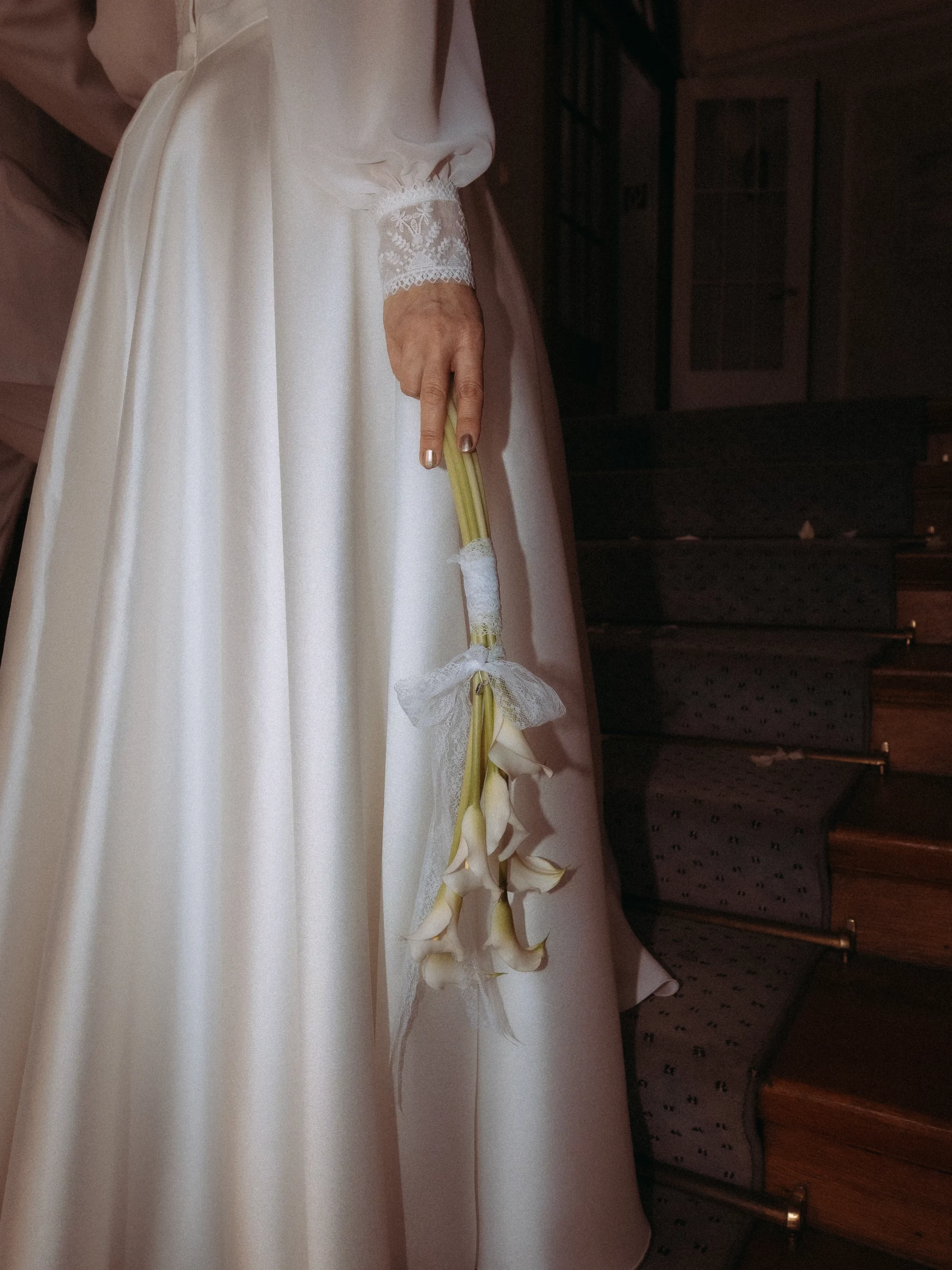 Eine Person in einem weißen Satinkleid steht auf einer Treppe und hält einen Strauß Calla-Lilien in der Hand, der mit einer weißen Schleife gebunden ist - die perfekte Inspiration für Hochzeitsfotos MV. Nur die untere Hälfte ihres Körpers ist sichtbar.
