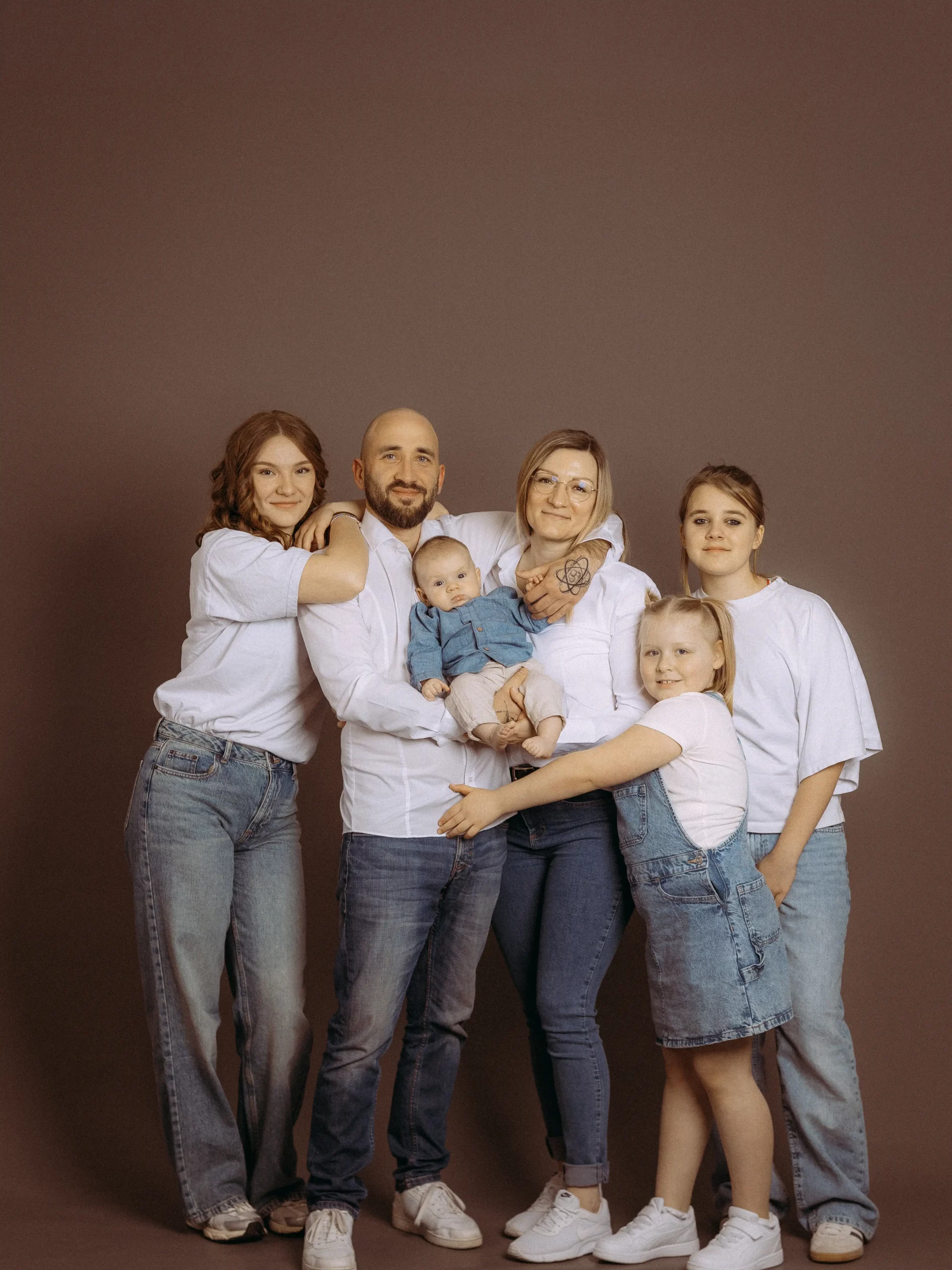 Familienfoto MV: Glückliche Familie mit Baby, zwei Töchtern und Eltern vor braunem Hintergrund. Alle tragen Jeans und weiße Oberteile. Familienfotograf in Schwerin.