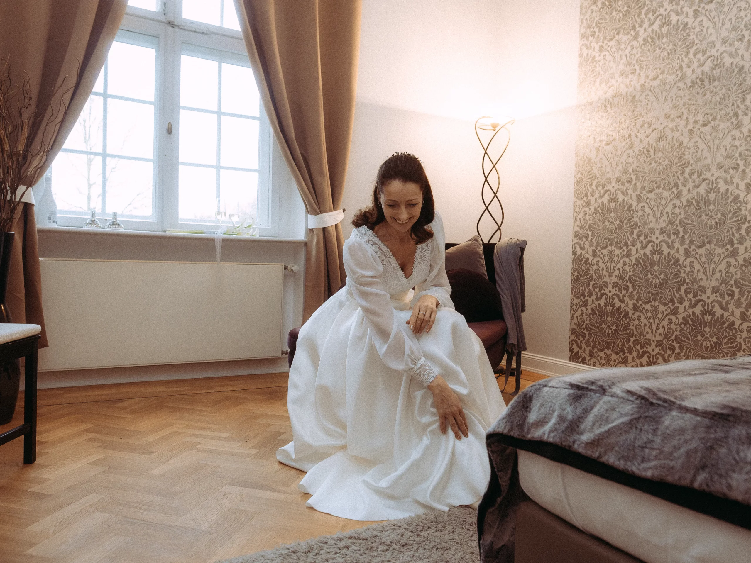 Eine Frau in einem langen weißen Kleid kniet auf dem Holzboden eines gemütlichen, warm beleuchteten Schlafzimmers und lächelt, während sie ihr Kleid berührt. Das von einem Hochzeitsfotograf MV aufgenommene Zimmer verfügt über große Fenster mit Vorhängen und gemusterten Tapeten.