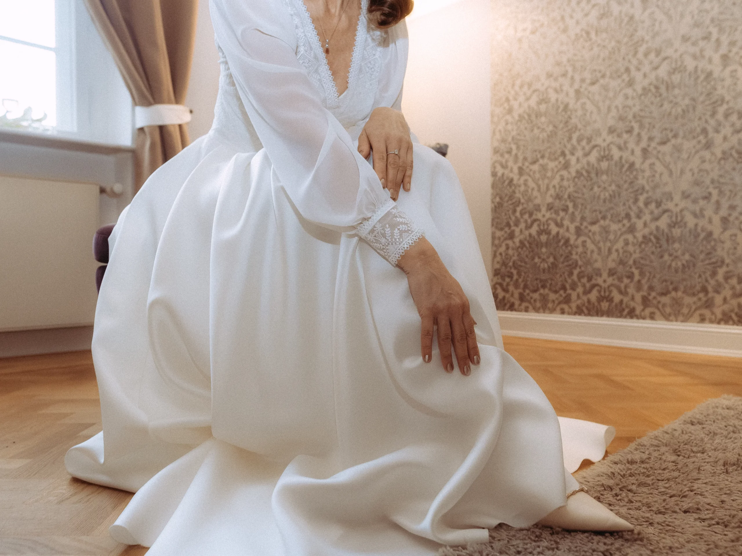Eine Person, die ein langärmeliges weißes Hochzeitskleid trägt, kniet auf einem Holzboden neben einem beigen Teppich, eine Hand auf ihrem Knie. Der Raum hat helle Wände und gemusterte Tapeten - perfekt für Hochzeitsfotos MV.