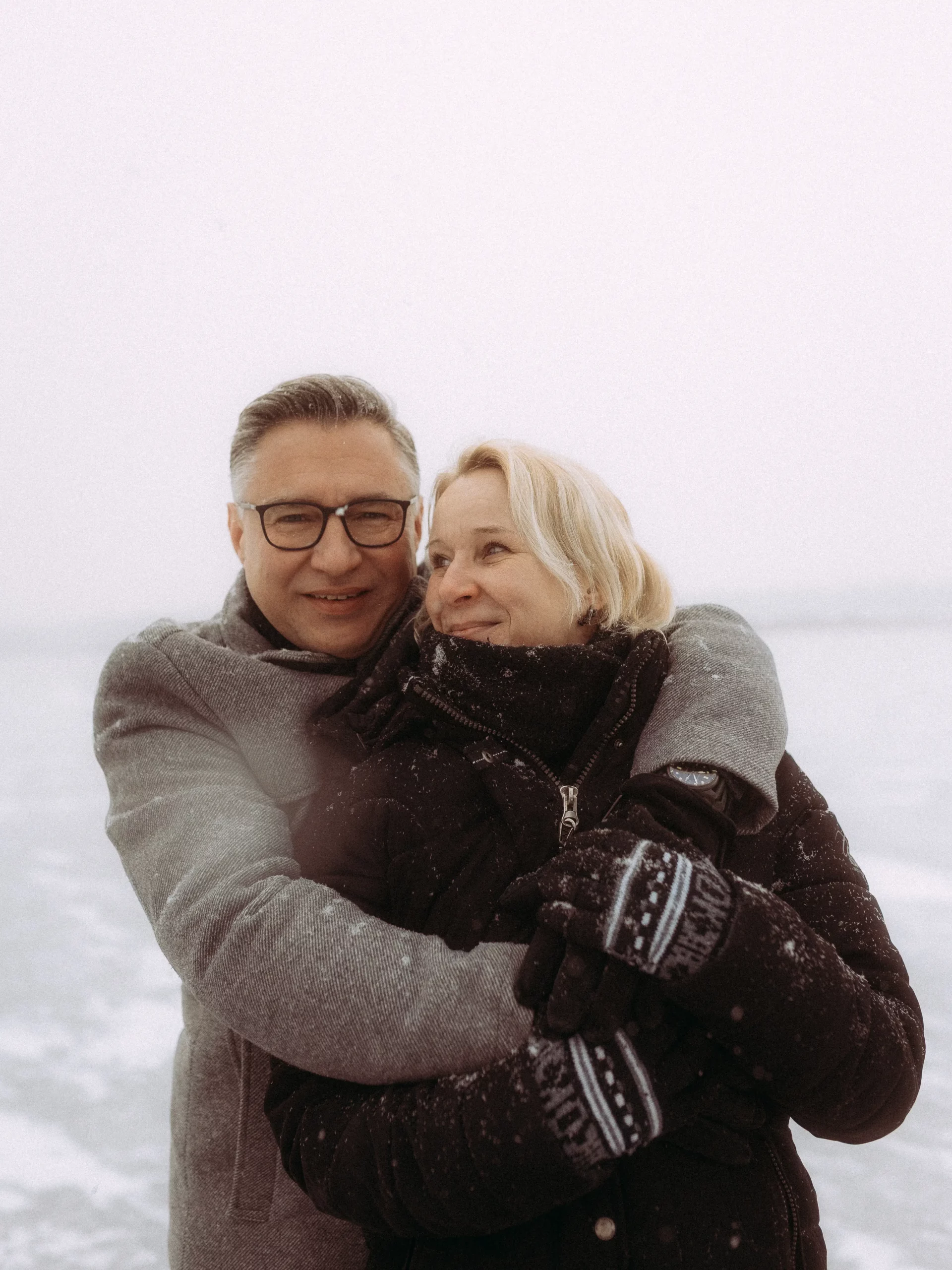 Ein Mann und eine Frau in Wintermänteln und Handschuhen umarmen sich und lächeln, während sie an einem verschneiten, bewölkten Tag draußen stehen, aufgenommen von Fotograf MV.