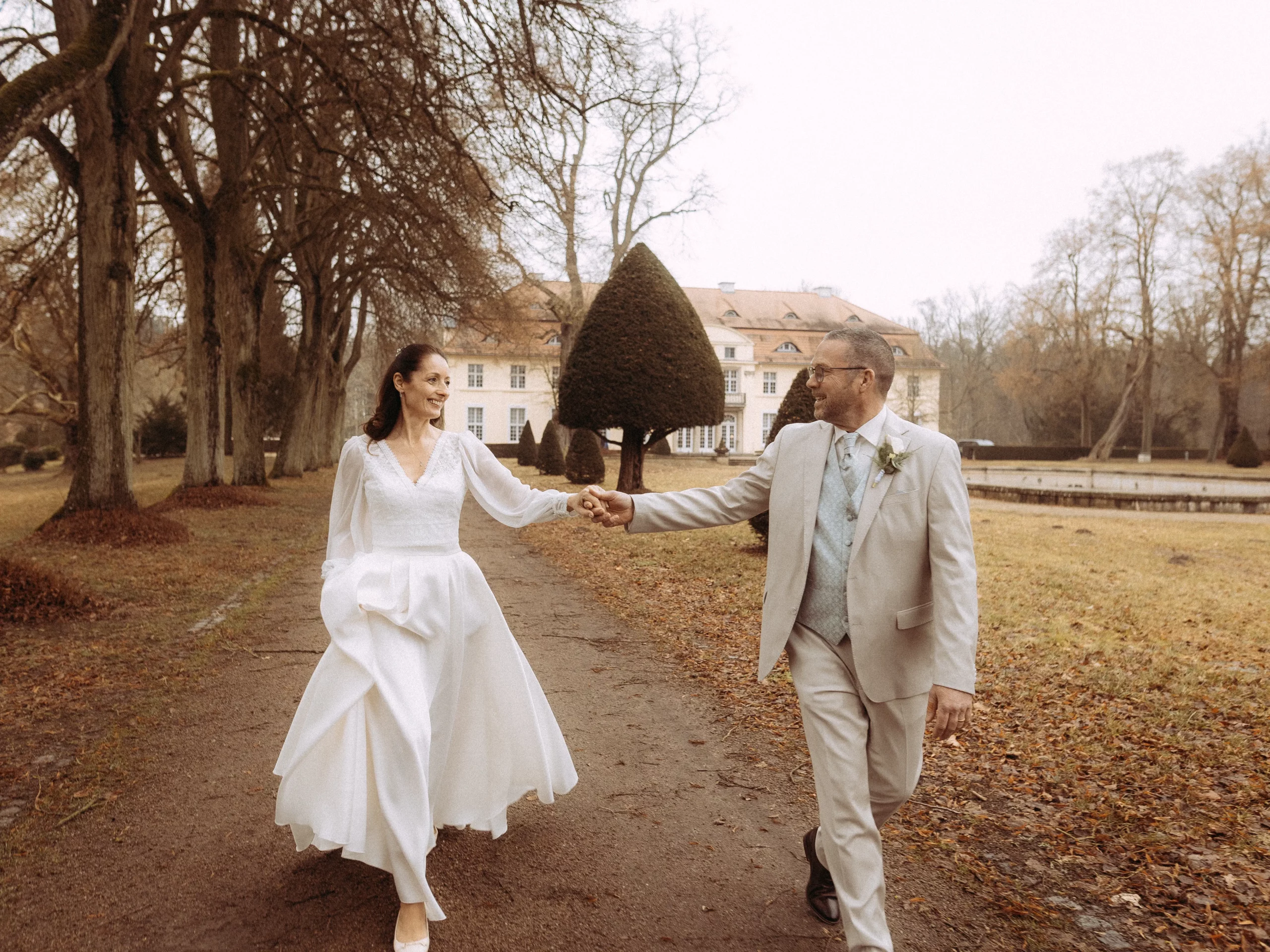 Brautpaar hält Händchen vor Schloss. Herbstliche Hochzeit im Park.