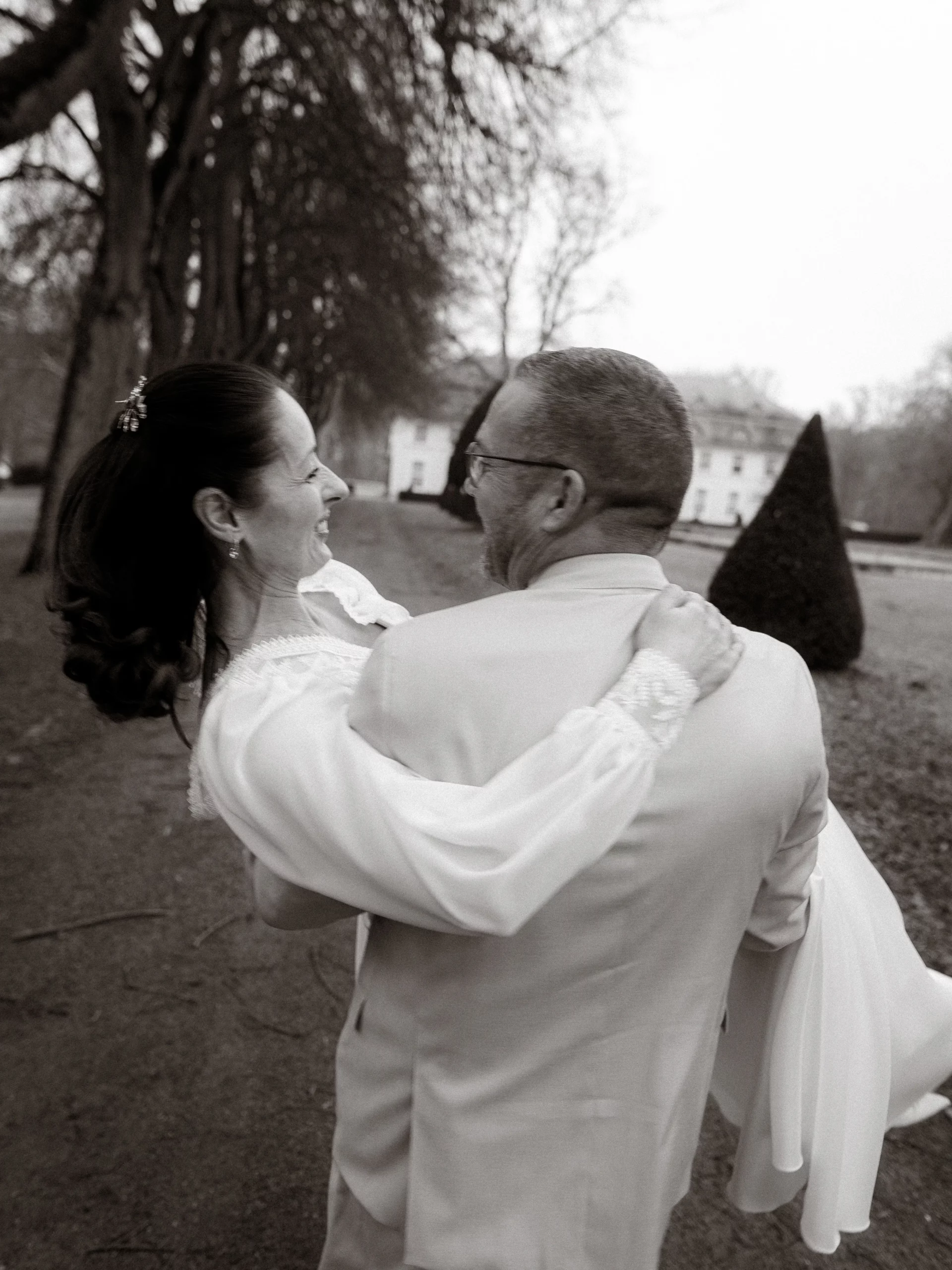 Brautpaar in Schwarzweißaufnahme. Der Bräutigam trägt die Braut im Arm, beide lachen.