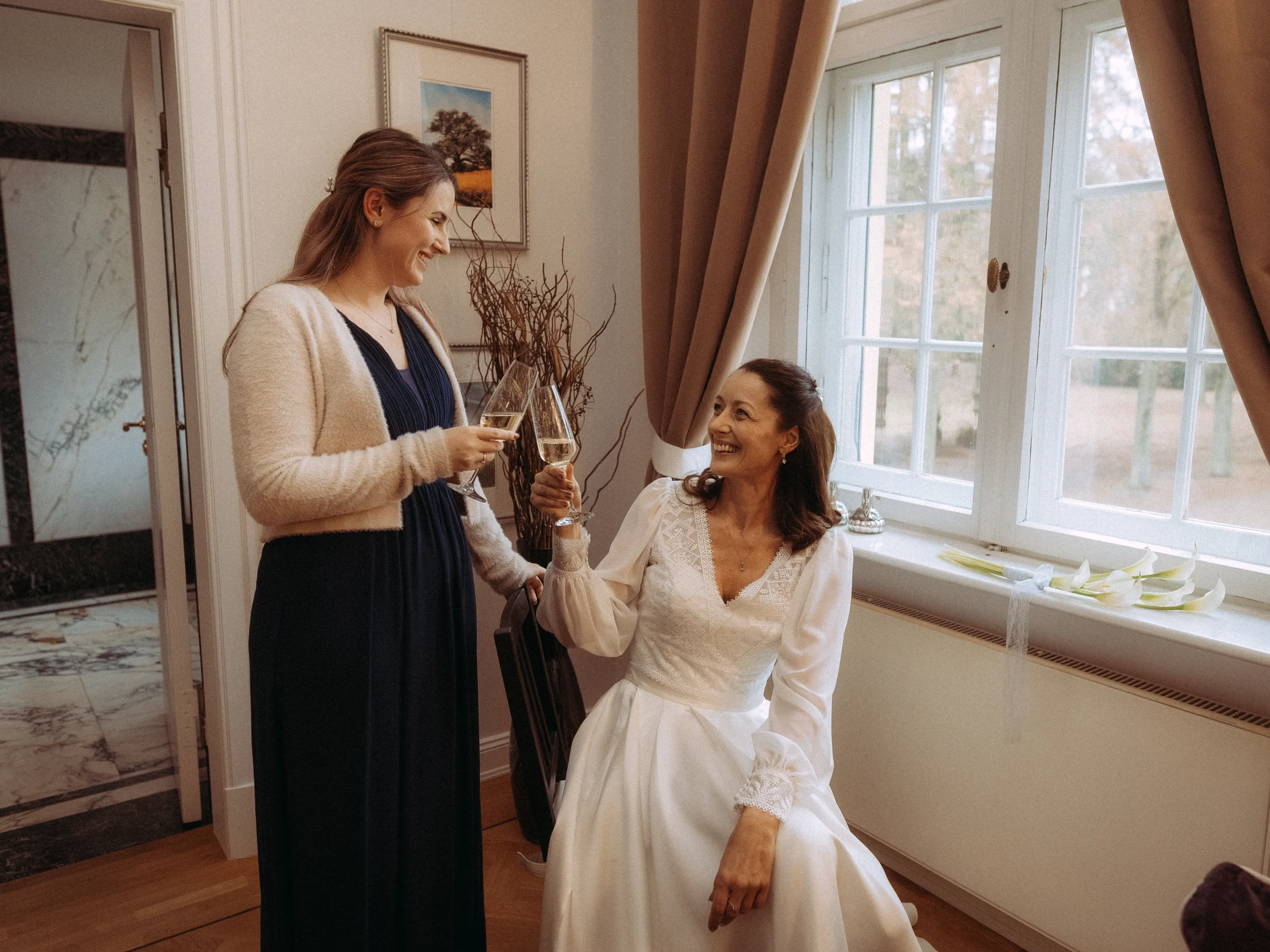 Zwei Frauen lächeln und stoßen mit Gläsern Wein in einem warm beleuchteten Raum an; eine in einem weißen Hochzeitskleid sitzt, während die andere in einem marineblauen Kleid neben ihr in der Nähe eines Fensters steht - aufgenommen von Hochzeitsfotograf MV für unvergessliche Hochzeitsfotos MV.