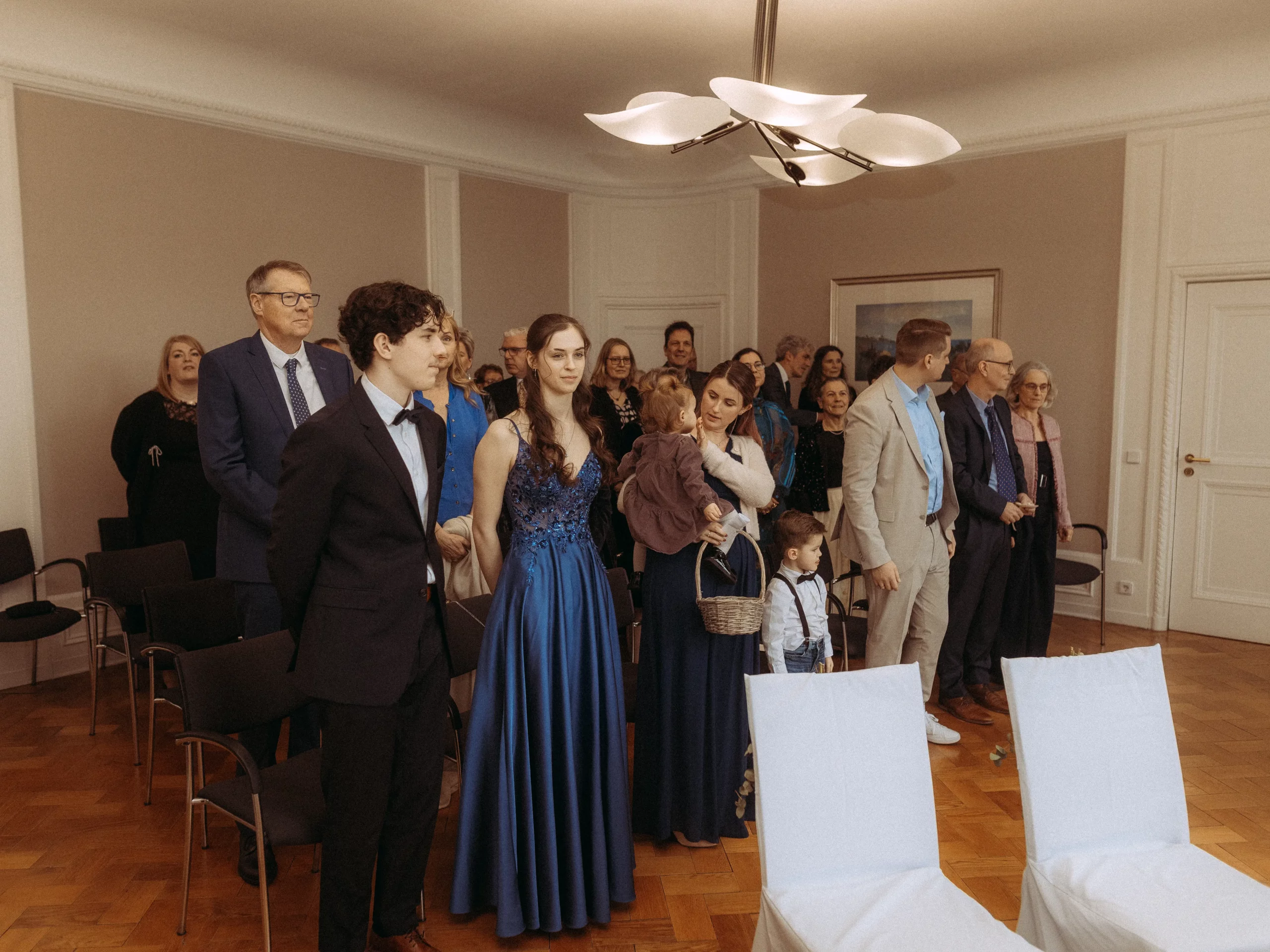 Eine Gruppe gut gekleideter Menschen, darunter auch Kinder, steht in einem formellen Raum mit leeren weißen Stühlen im Vordergrund und wartet auf eine Zeremonie - perfekt für Hochzeitsfotograf MV, um unvergessliche Hochzeitsfotos MV festzuhalten.