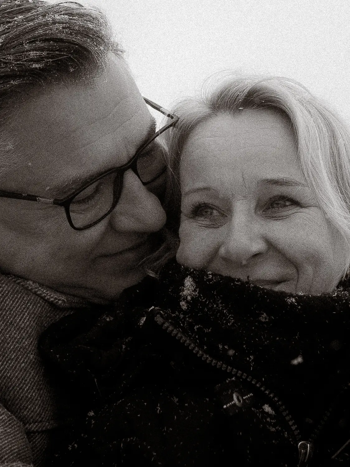 Ein Schwarz-Weiß-Foto in Nahaufnahme von einem lächelnden Paar in warmer Kleidung. Der Mann, der eine Brille trägt, lehnt sich liebevoll an die Frau, während sie einen verschneiten Moment im Freien genießen - perfekt für Hochzeitsfotos MV.