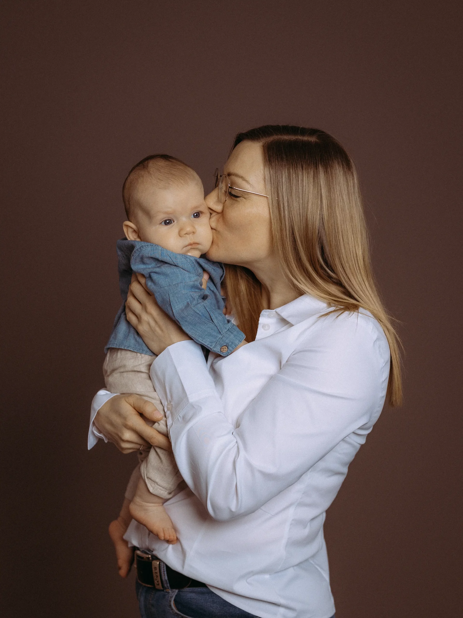 Liebevolle Mutter hält ihr Baby im Arm und gibt ihm einen Kuss. Familienfoto in Schwerin zeigt Zuneigung vor braunem Hintergrund, beide tragen helle Kleidung. Fotograf aus Schwerin.