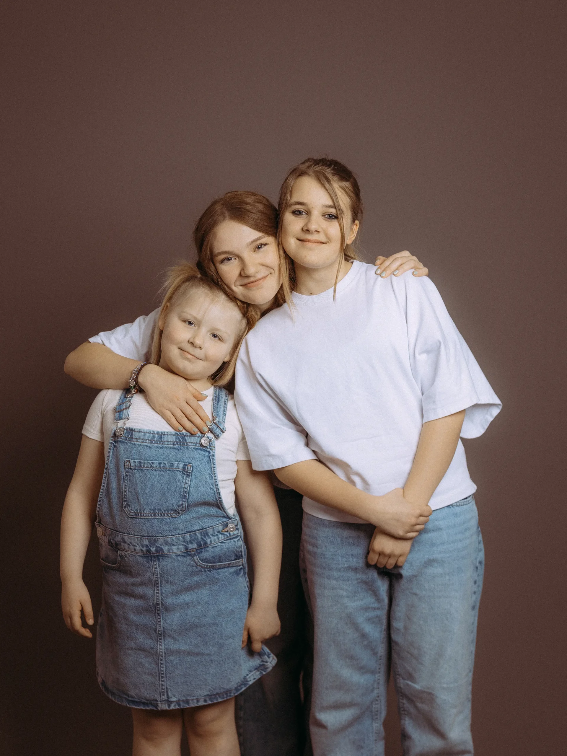 Drei Schwestern umarmen sich herzlich vor neutralem Hintergrund. Das jüngste Mädchen trägt ein Jeanskleid, die älteren Schwestern weiße TShirts und Jeans. Familienfotos MV.