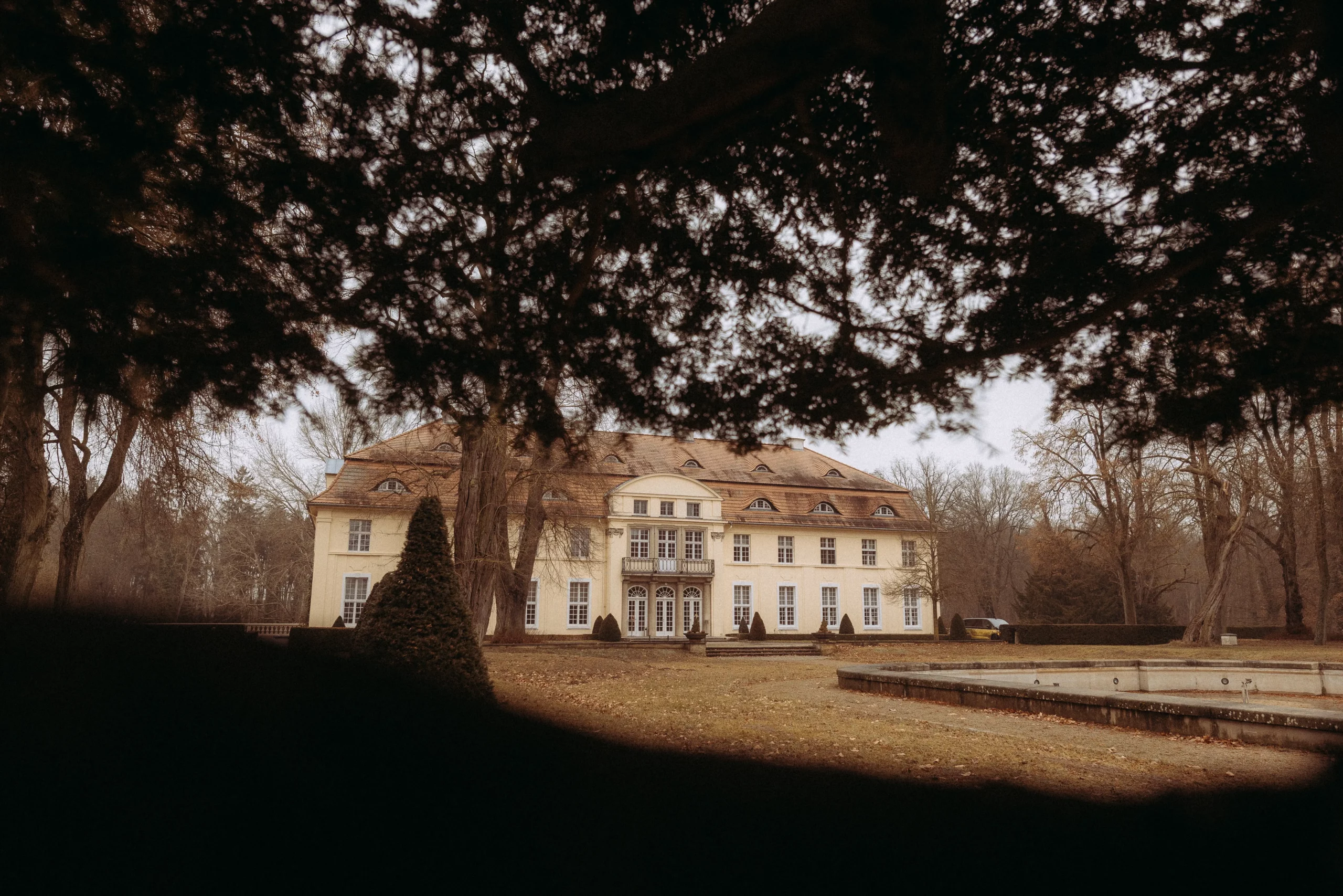 Ein großes, elegantes Herrenhaus mit vielen Fenstern und einem spitzen Dach steht inmitten von blattlosen Bäumen, die durch eine Lücke im dichten Laubwerk betrachtet werden. Perfekt für einen Hochzeitsfotograf MV oder Familienfotograf MV, um atemberaubende winterliche Szenen mit braunem Gras und einem leeren Springbrunnen einzufangen.