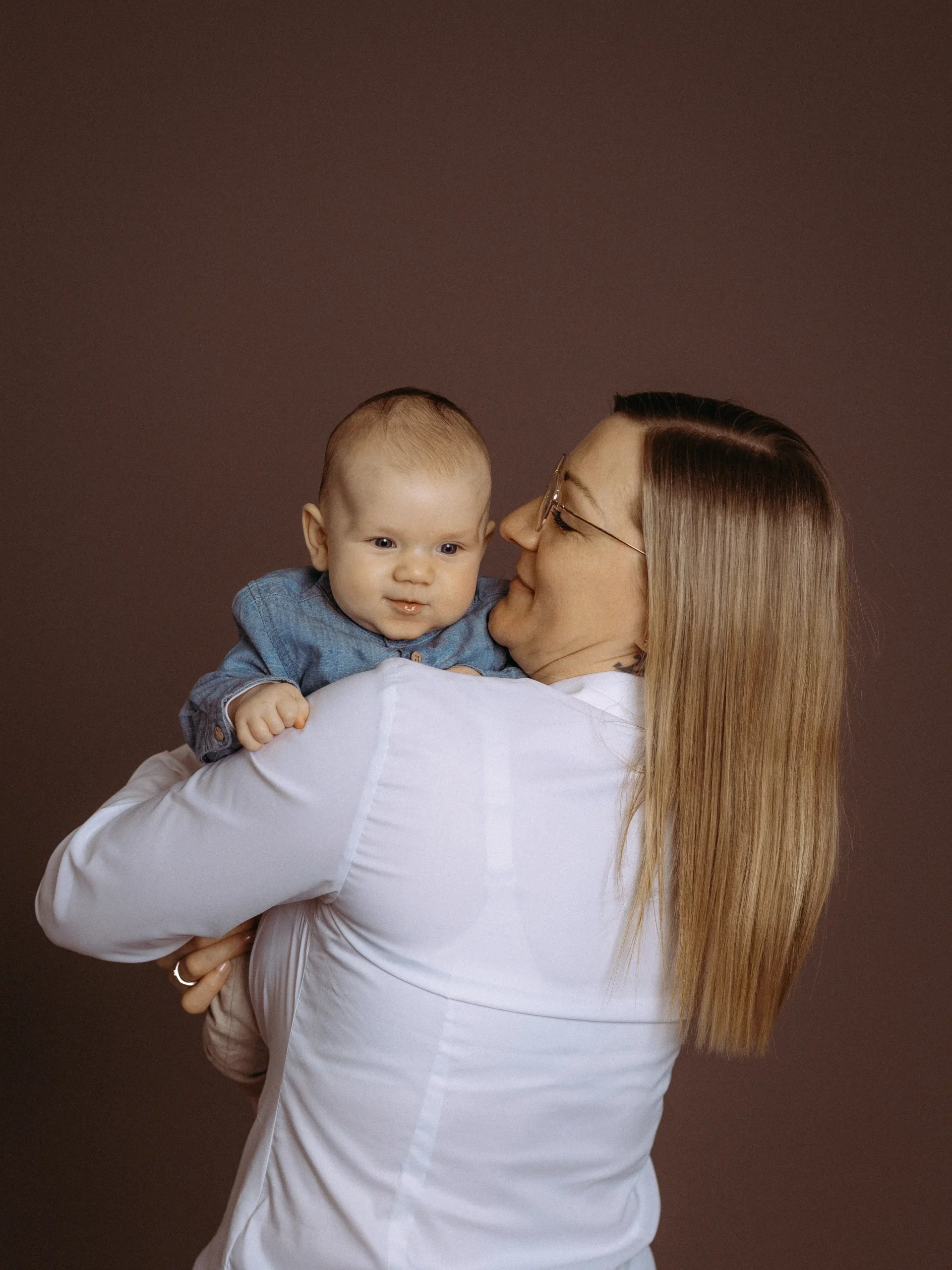 Eine liebevolle Mutter hält ihr Baby im Arm, beide lächeln. Familienfotos MV. Die Mutter trägt eine weiße Bluse und das Baby ein blaues Hemd. Fotograf in MV.