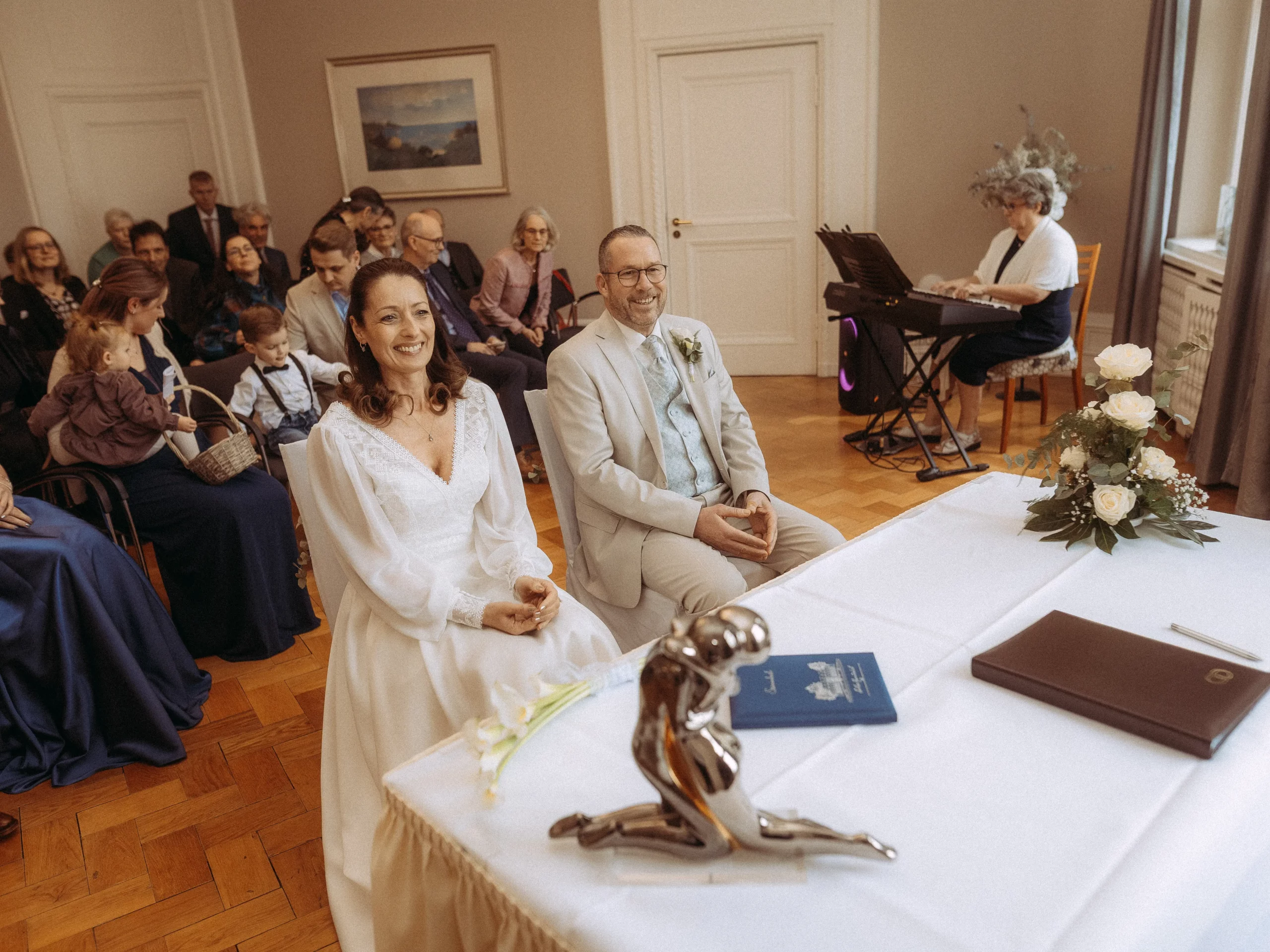 Braut und Bräutigam sitzen lächelnd an einem Tisch während ihrer Hochzeitszeremonie in einem hellen Raum, mit Gästen hinter ihnen und einer Frau, die Keyboard spielt. Ein Blumenstrauß und eine Skulptur stehen auf dem Tisch - perfekt für Hochzeitsfotos MV.
