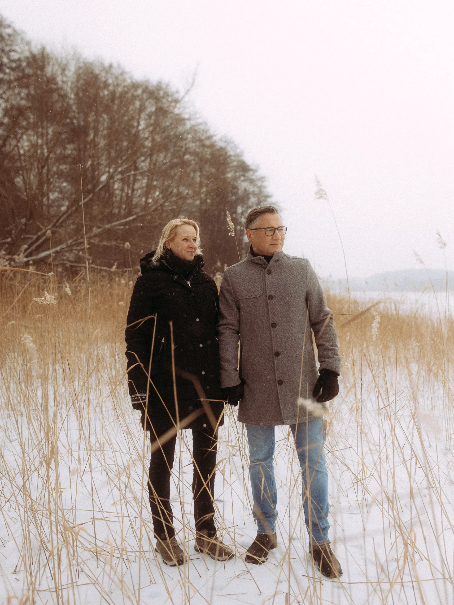 Ein Paar in Wintermänteln und Handschuhen steht dicht beieinander auf einem verschneiten Feld mit hohem, trockenem Gras, Bäumen im Hintergrund und einem blass bewölkten Himmel - perfekt für natürliche Hochzeitsfotos MV.