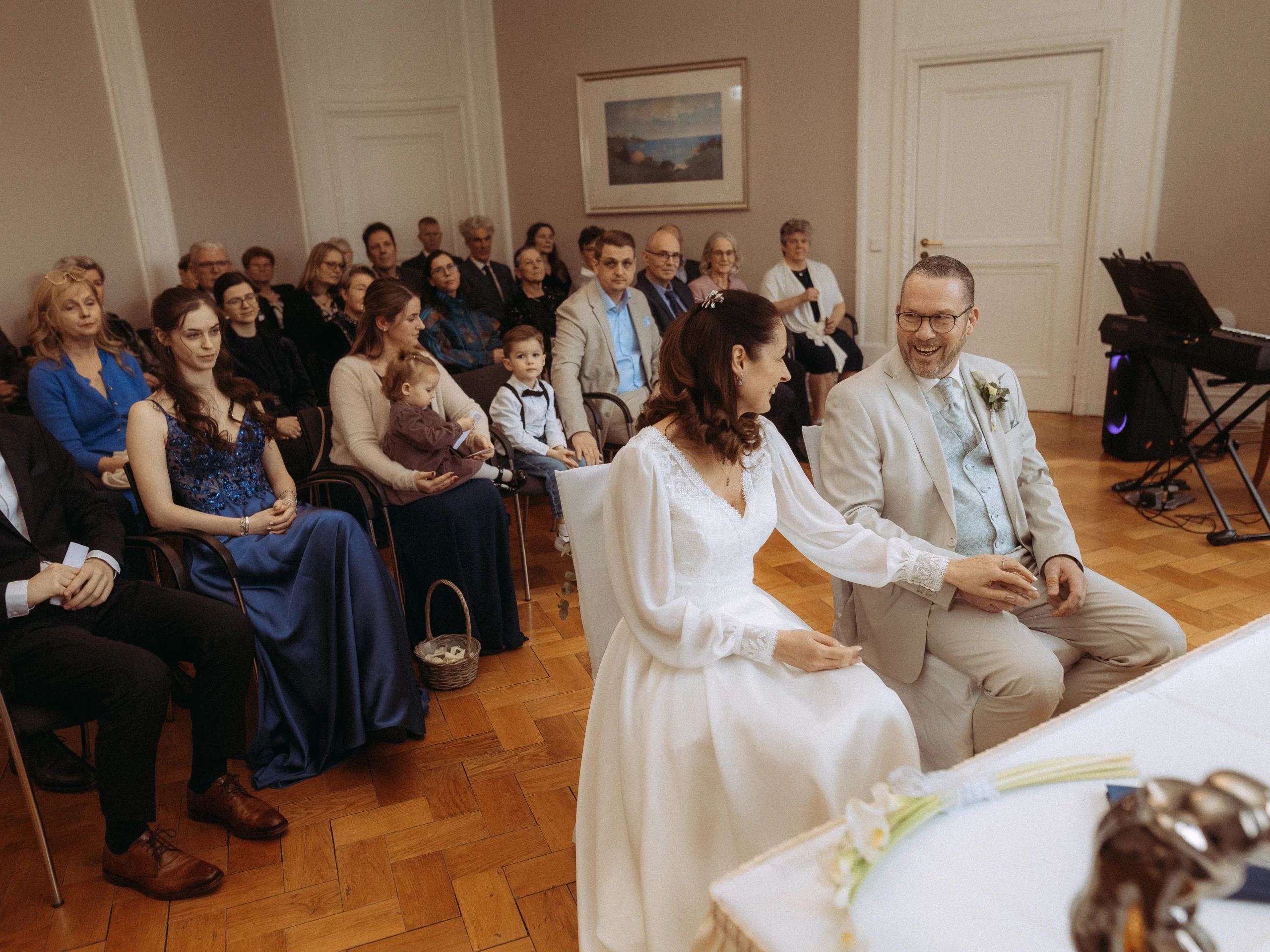 Braut und Bräutigam sitzen sich während ihrer Hochzeitszeremonie lächelnd gegenüber, umgeben von fröhlichen Gästen jeden Alters. Der intime Moment wird von einem Hochzeitsfotograf MV wunderschön festgehalten und schafft bleibende Erinnerungen für das Paar.
