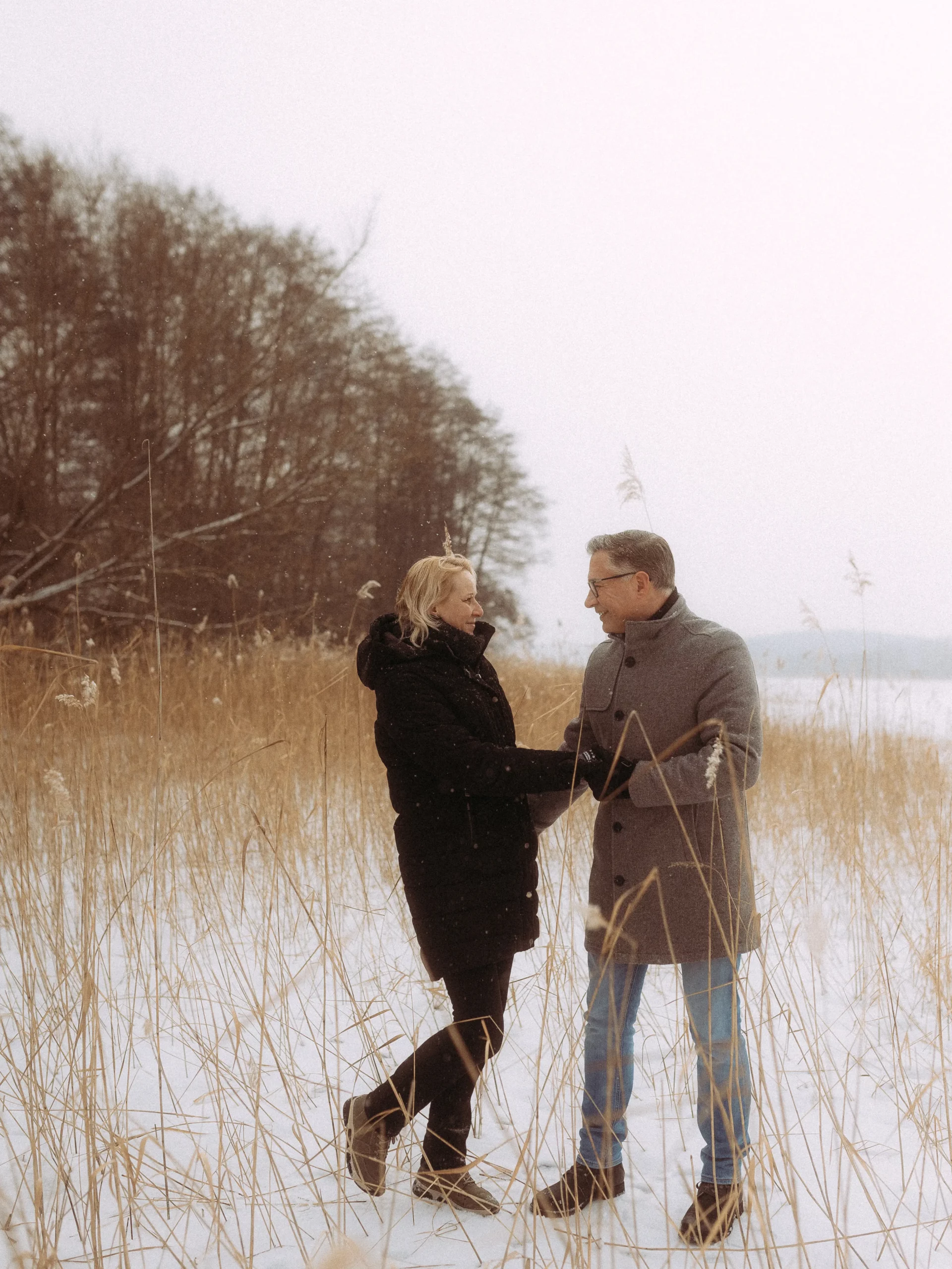 Ein in Wintermäntel gekleidetes Paar steht händchenhaltend auf einem verschneiten Feld mit hohem, trockenem Gras und kahlen Bäumen im Hintergrund und teilt einen warmen Moment an einem kalten Tag - aufgenommen von Hochzeitsfotograf MV.