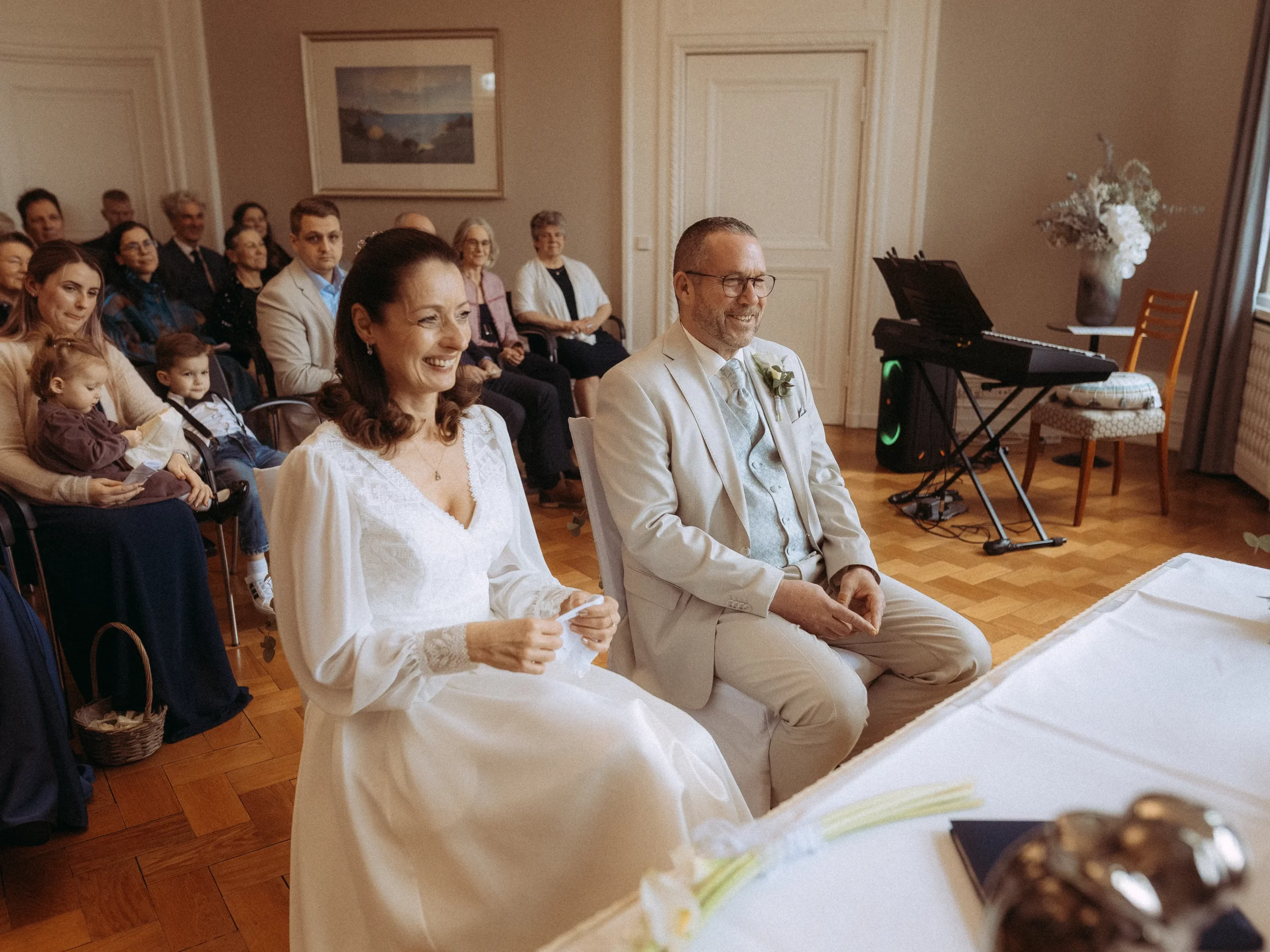 Eine Braut und ein Bräutigam sitzen während ihrer Hochzeitszeremonie lächelnd an einem Tisch und blicken auf einen Zeremonienmeister. Die Gäste sitzen hinter ihnen und schauen aufmerksam in einem warm beleuchteten Raum mit Holzboden zu - aufgenommen von Hochzeitsfotograf MV.