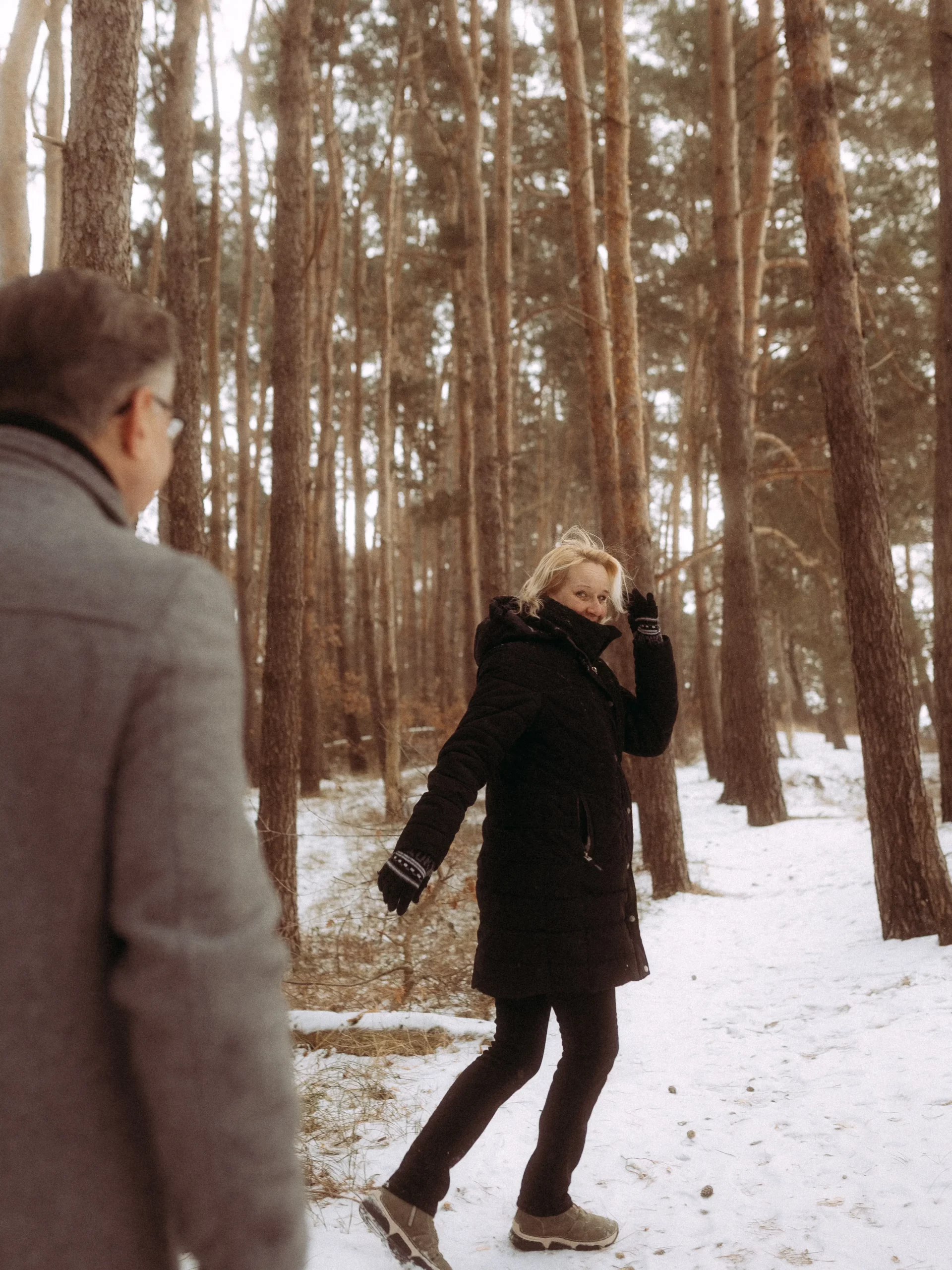 Eine Frau in einem schwarzen Mantel lächelt und winkt, während sie durch einen verschneiten Wald mit hohen Kiefern geht, während ein Mann in einem grauen Mantel hinter ihr herläuft. Dieser von Fotograf MV aufgenommene Moment strahlt Winterfreude aus.