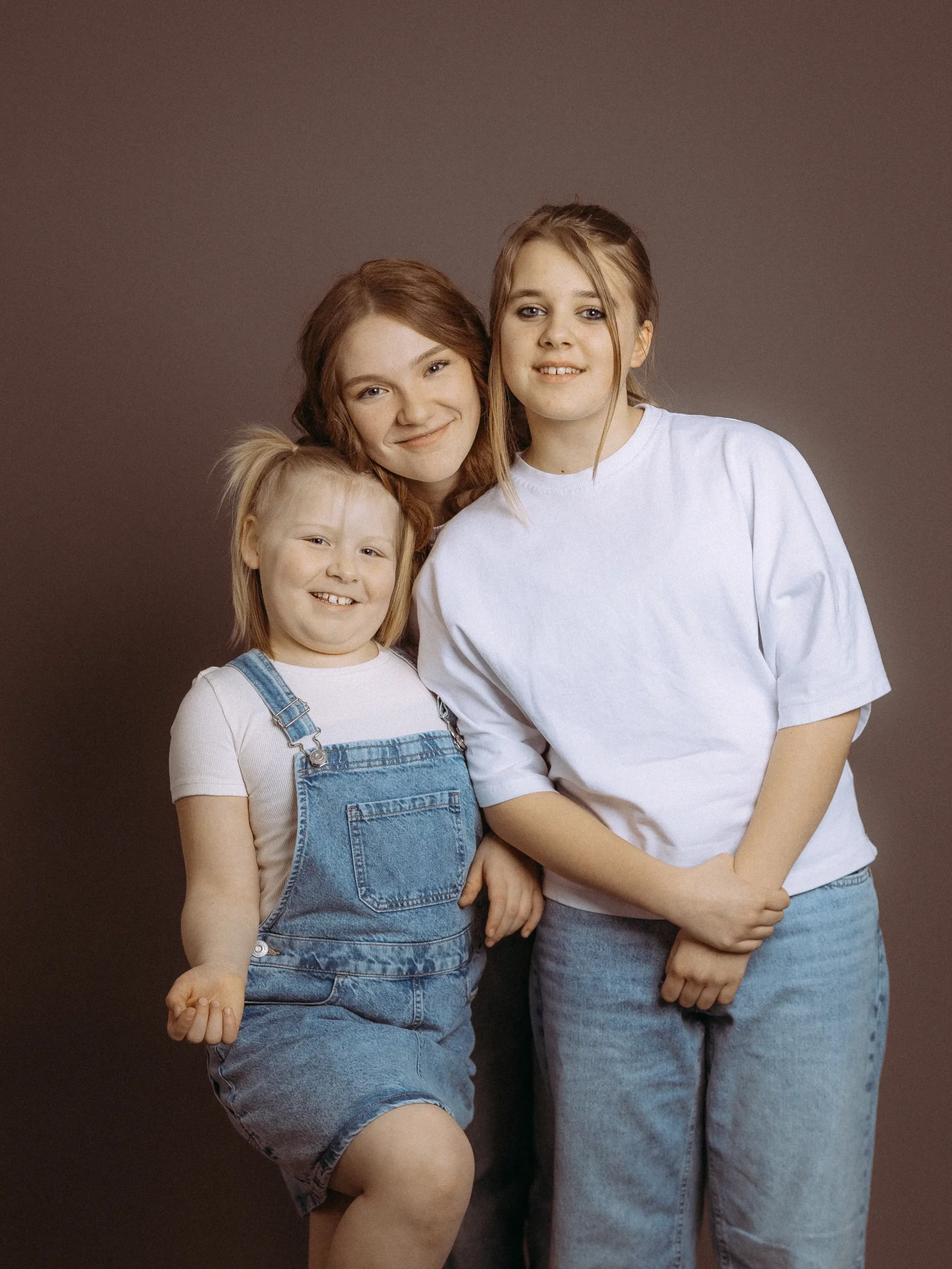 Drei lächelnde Schwestern posieren vor grauem Hintergrund. Die Jüngste trägt Jeanslatzhose, die Älteren weiße TShirts und Jeans. Familienfotos MV vom Fotograf aus Schwerin.