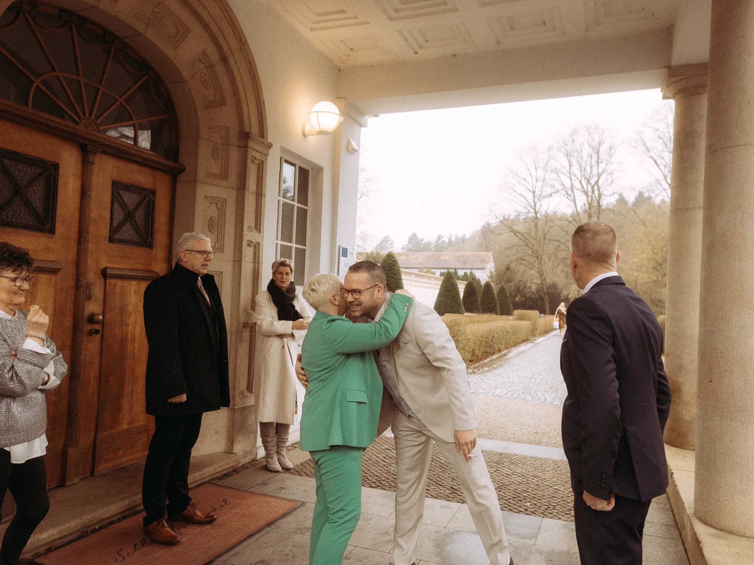 Eine Person in einem grünen Anzug umarmt und küsst jemanden in einem hellen Anzug vor einem Gebäude, während andere freudig zusehen - eingefangen von Hochzeitsfotos MV inmitten von Bäumen und einem Kopfsteinpflasterweg, der die Wärme einer Feier heraufbeschwört.