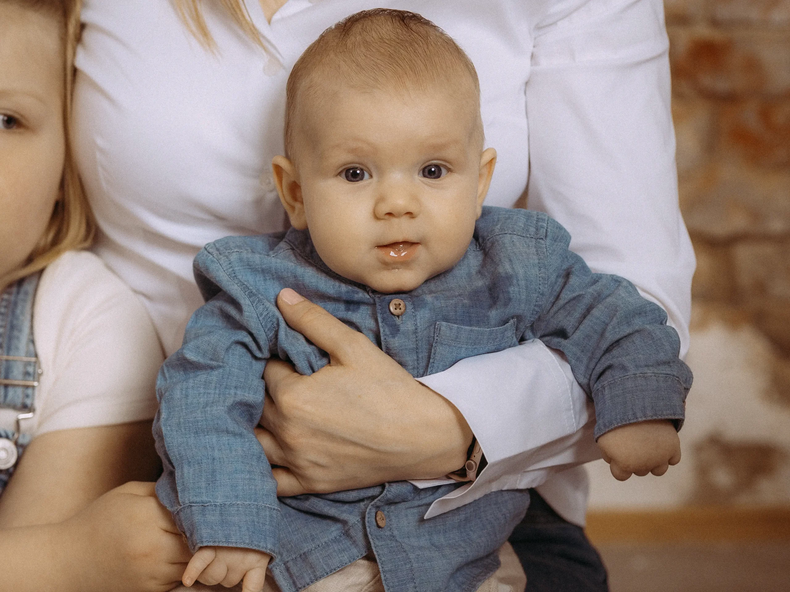 Ein bezauberndes Baby in den Armen einer Frau, trägt ein blaues Hemd. Familienfotos MV. Ein Geschwisterchen ist auch im Bild. Fotograf in MV.