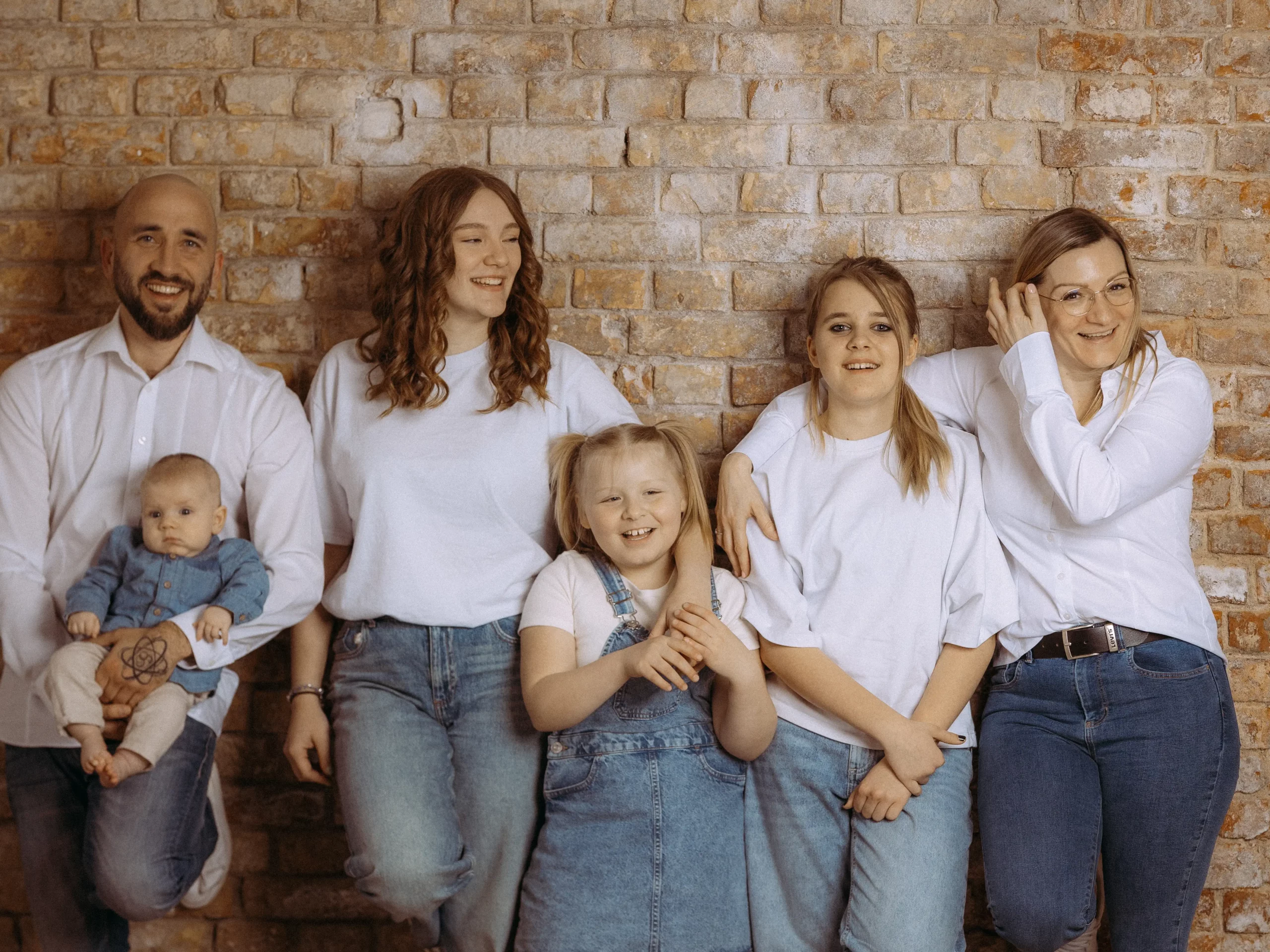 Familienfotos MV: Glückliche Familie vor Backsteinmauer. Vater hält Baby, Mädchen im Jeanskleid lächelt, Mutter mit Brille. Freundliche Atmosphäre, natürliches Licht. Fotograf in MV.