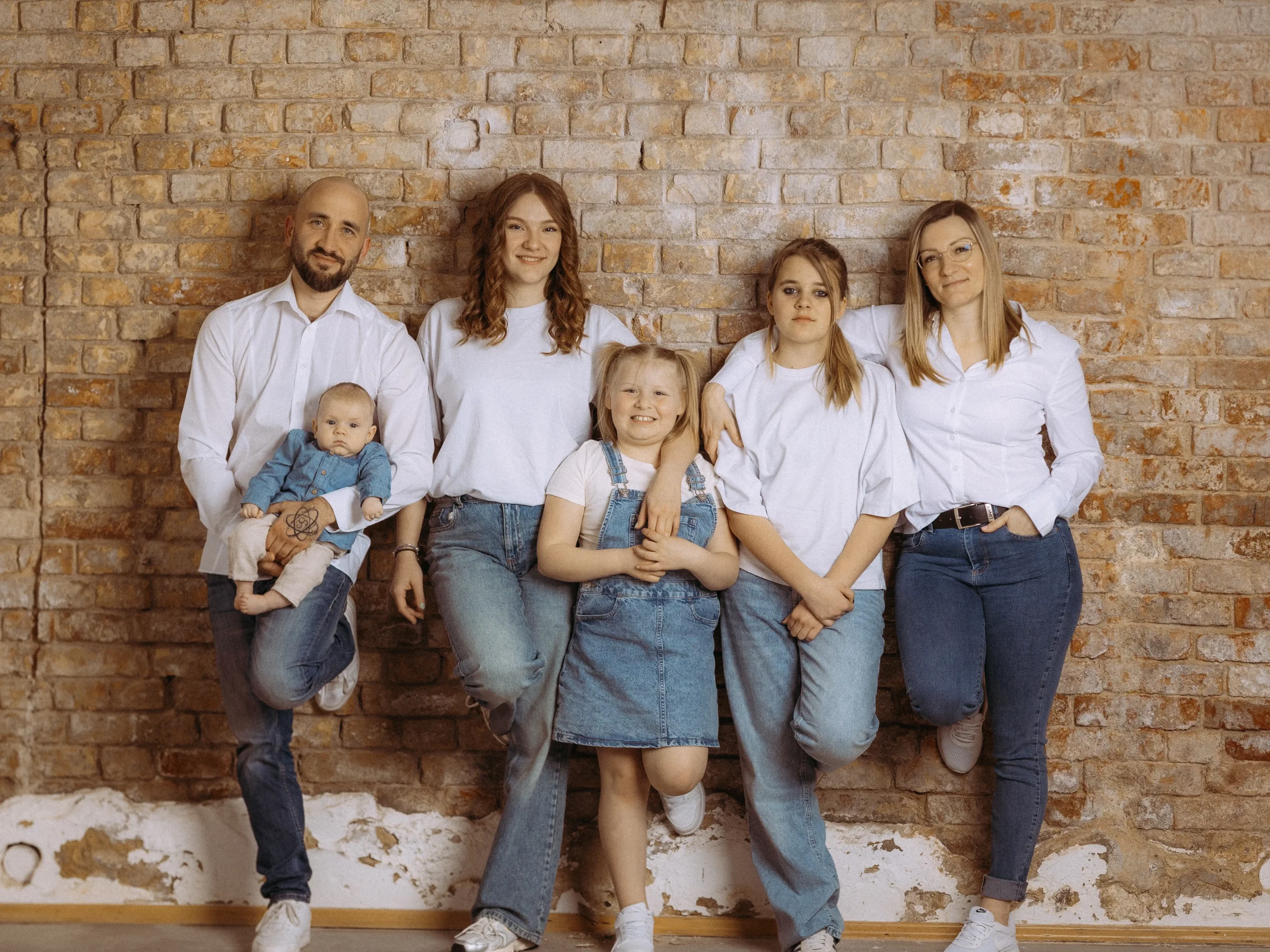 Familienfoto MV: Familie lehnt an einer Backsteinmauer. Der Vater hält ein Baby, während die Töchter und die Mutter lächeln. Fotograf aus Schwerin hat diese herzliche Szene eingefangen.