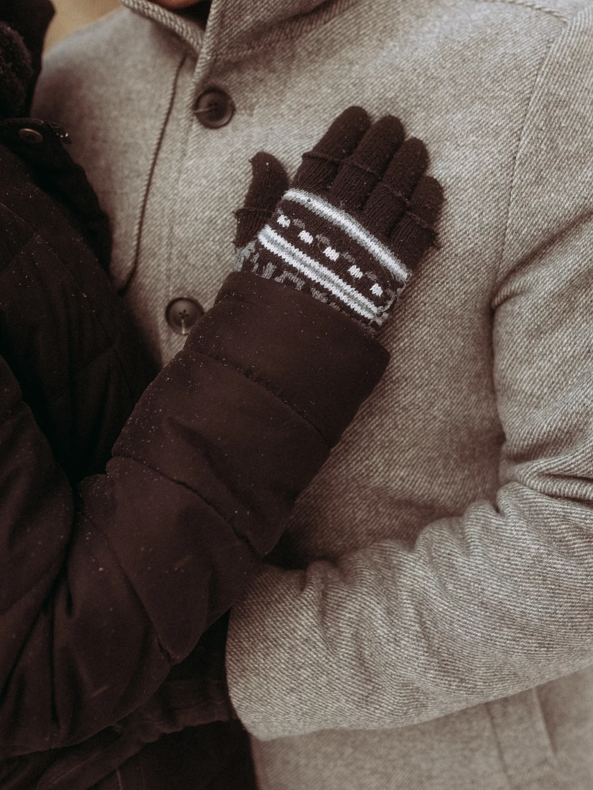 Eine Person, die gemusterte schwarze Handschuhe und einen dunklen Mantel trägt, legt ihre Hand auf die Brust einer anderen Person in einem hellgrauen Mantel mit Knöpfen, aufgenommen von Familienfotograf MV.
