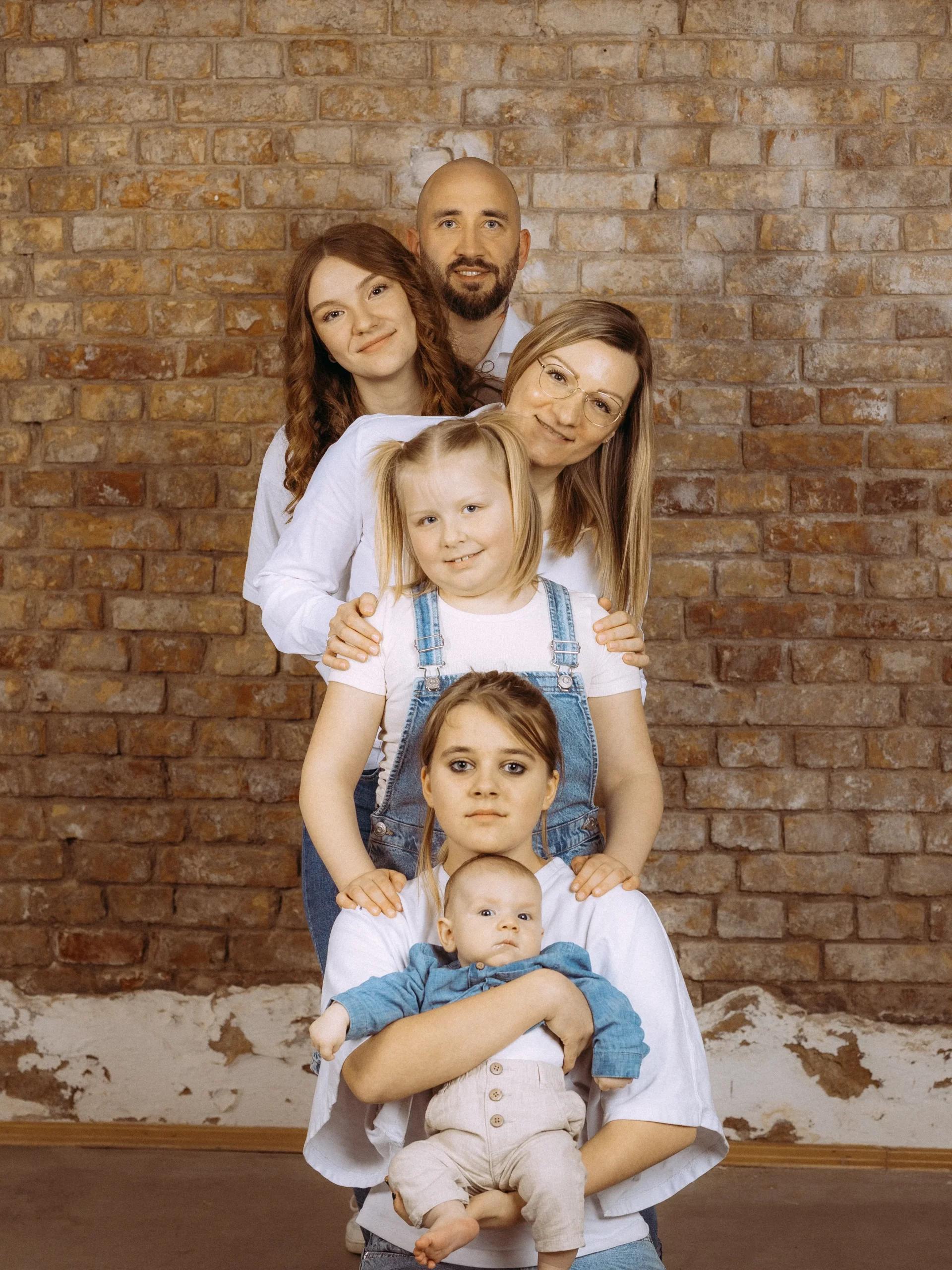 Familienfoto MV: Fünfköpfige Familie vor Backsteinmauer. Mutter hält Baby, darüber drei Mädchen und Vater. Freundliche, harmonische Familienfotos MV vom Fotograf aus Schwerin.