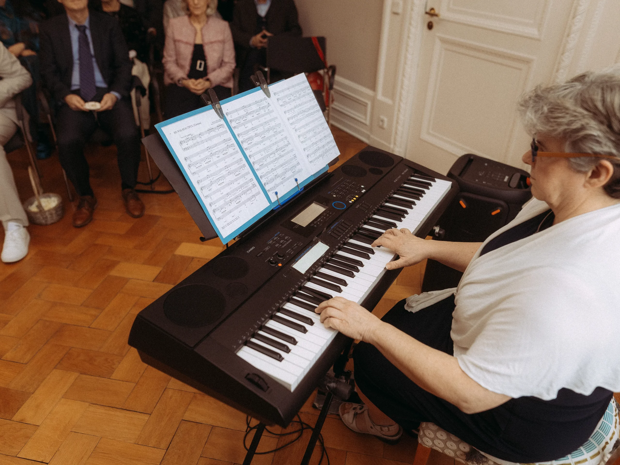 Eine Frau spielt auf einem elektrischen Keyboard mit Notenblättern, während mehrere sitzende Personen in einem hellen Raum mit Holzboden zusehen. Hochzeitsfotograf MV fängt diese besonderen Momente in einer warmen, einladenden Umgebung ein.