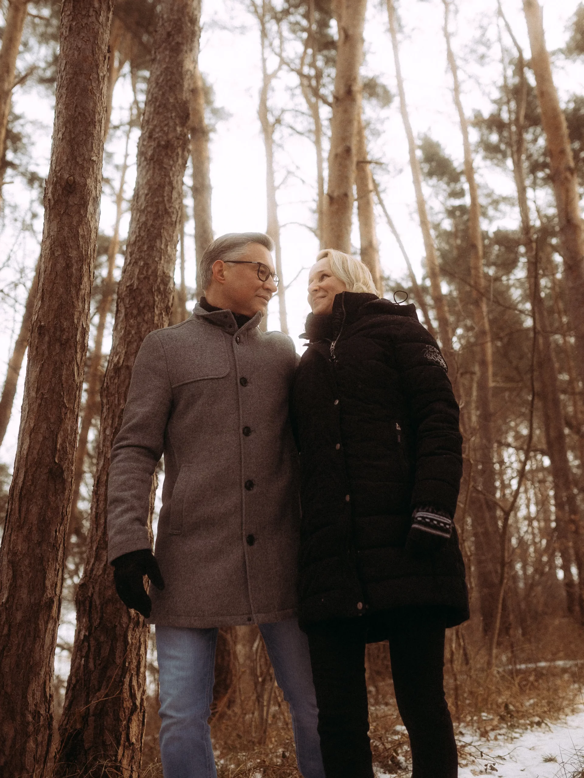 Ein Mann und eine Frau in Wintermänteln stehen eng beieinander und lächeln sich in einem verschneiten Wald mit hohen Bäumen um sie herum an - eingefangen von Fotograf MV für authentische, unvergessliche Momente.