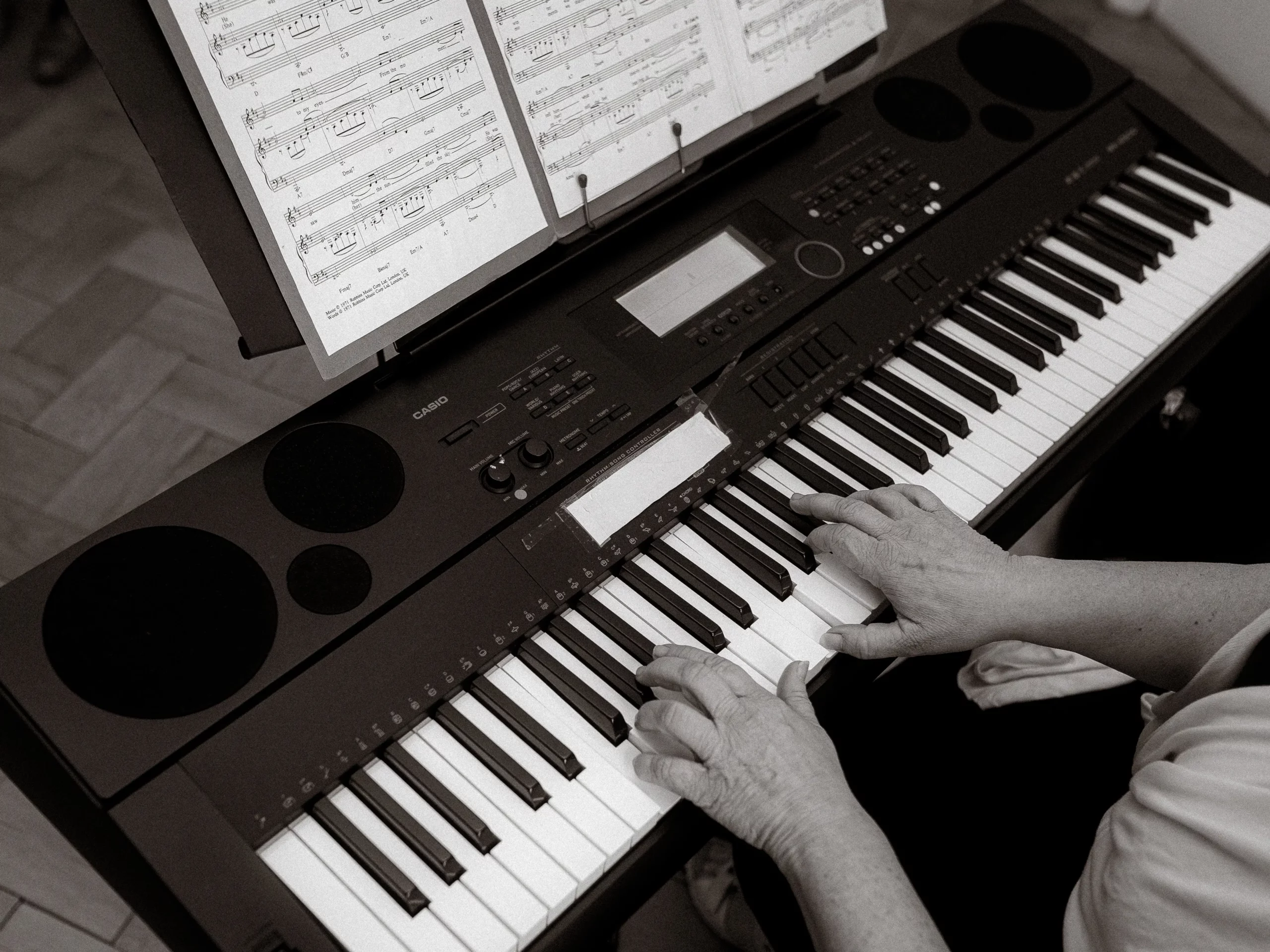 Eine Person spielt mit beiden Händen auf einem elektronischen Keyboard und liest dabei die Noten auf einem Ständer über den Tasten. Der von Fotograf MV in Schwarz-Weiß aufgenommene Moment unterstreicht den künstlerischen Fokus und die musikalische Leidenschaft.