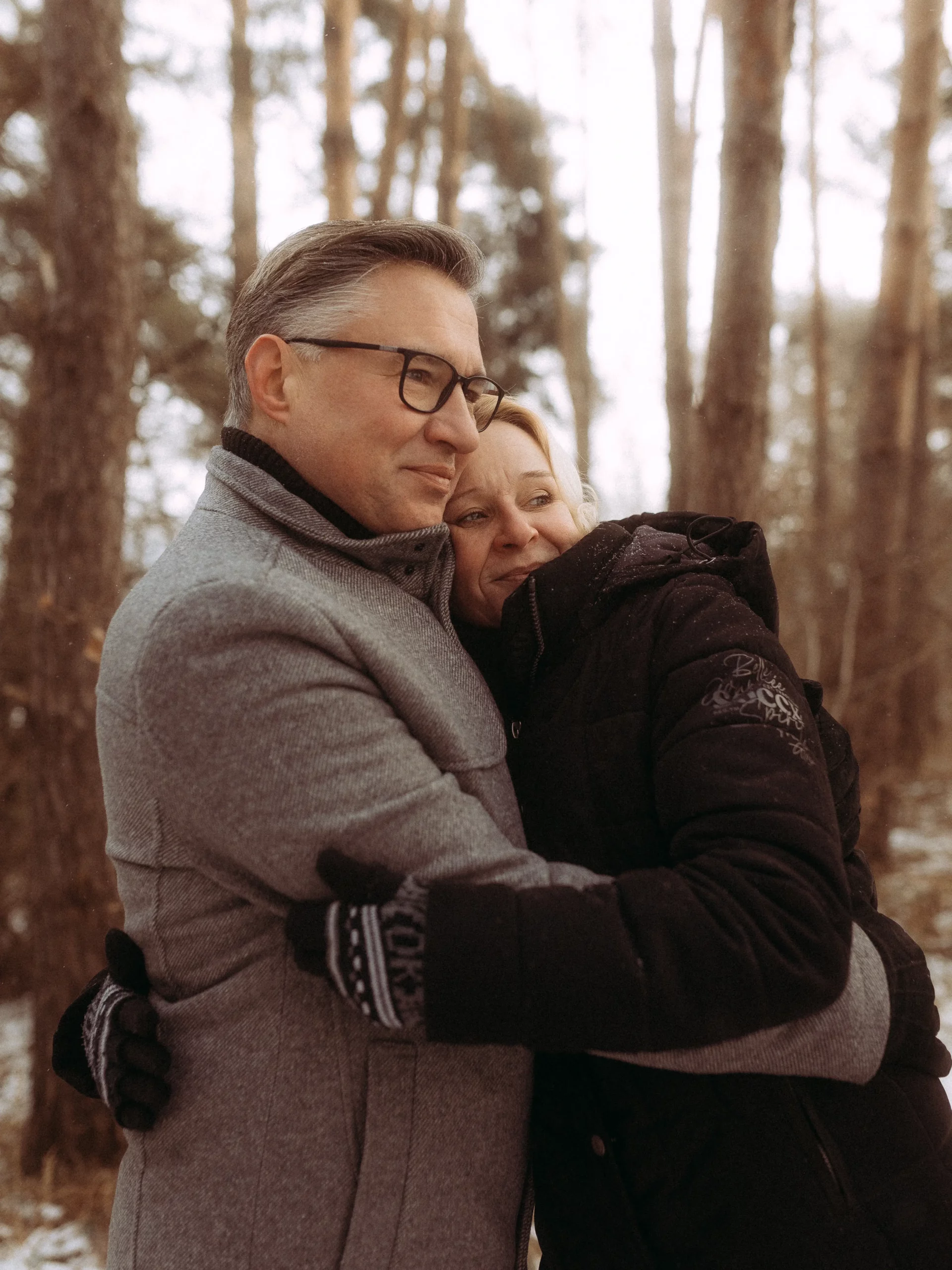 Ein Paar mittleren Alters in Wintermänteln und Handschuhen umarmt sich herzlich in einem verschneiten Wald und sieht zufrieden zwischen hohen Bäumen aus - aufgenommen von Hochzeitsfotograf MV für unvergessliche Hochzeitsfotos MV.