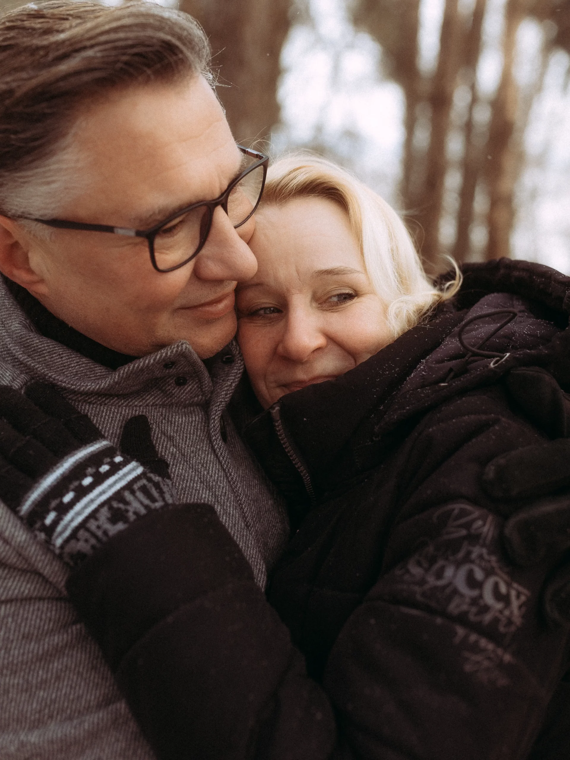 Ein lächelndes, winterlich gekleidetes Paar umarmt sich im Freien zwischen verschwommenen Bäumen. Die Frau schmiegt sich an den Mann, beide wirken warm und zufrieden - ein herzlicher Moment, eingefangen von Familienfotograf MV.