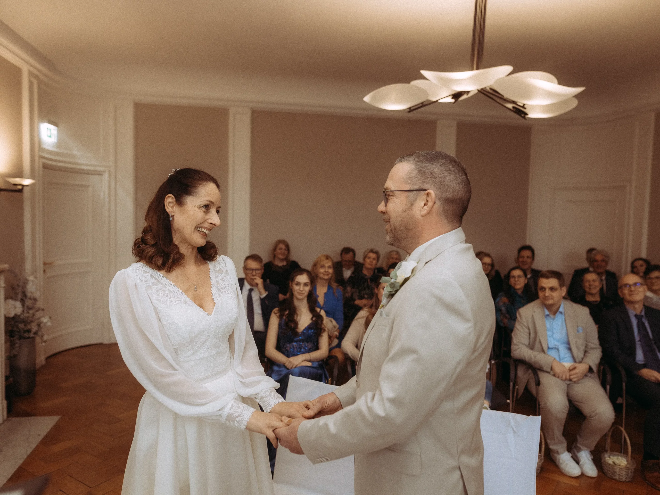 Eine Braut und ein Bräutigam stehen sich gegenüber, halten sich an den Händen und lächeln während ihrer Hochzeitszeremonie in einem Raum voller sitzender Gäste. Hochzeitsfotograf MV fängt die fröhliche und intime Atmosphäre wunderschön ein.