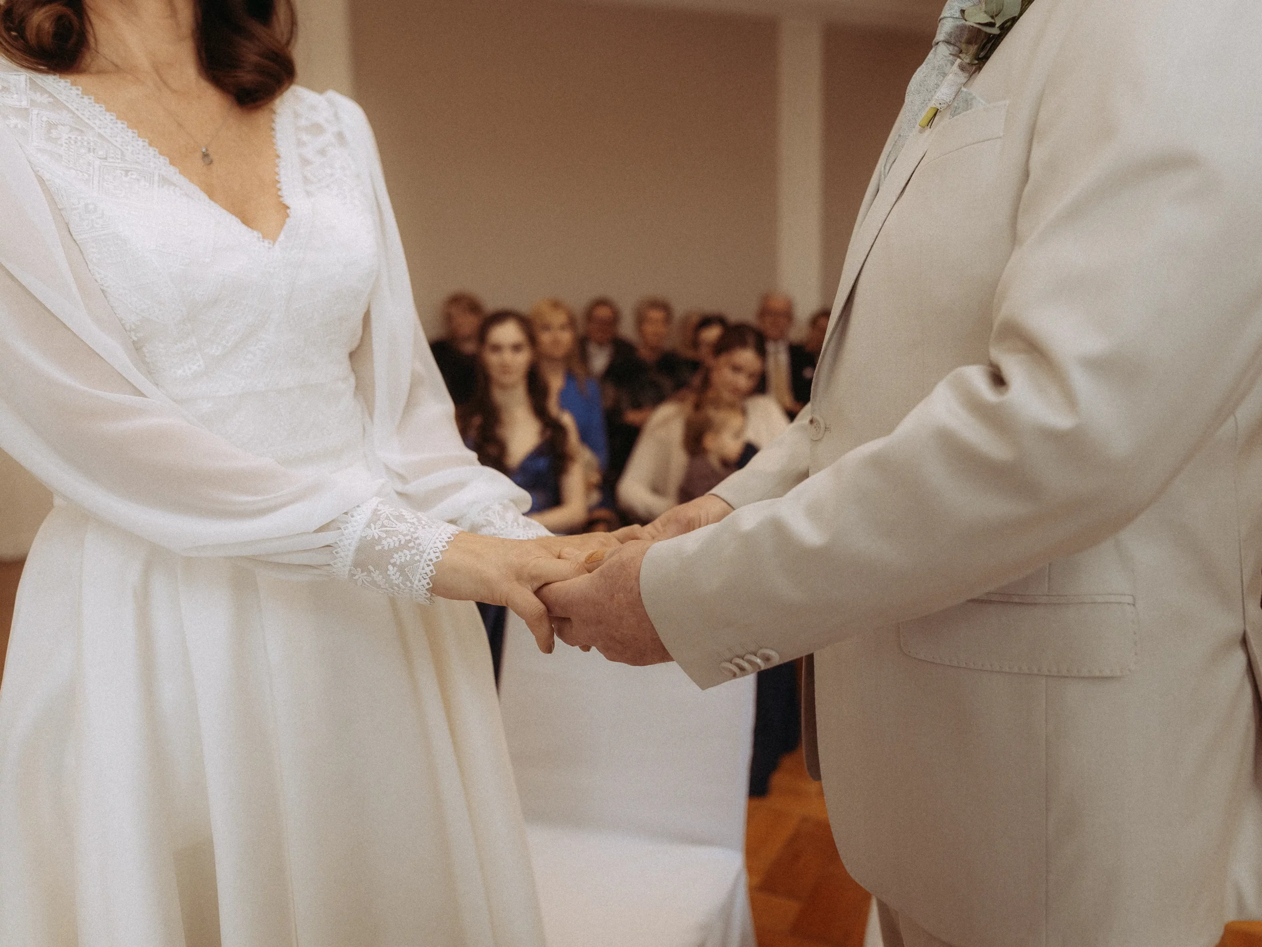 Eine Braut und ein Bräutigam halten sich während ihrer Hochzeitszeremonie an den Händen, gekleidet in ein weißes Kleid und einen hellen Anzug, mit sitzenden Gästen im verschwommenen Hintergrund. Hochzeitsfotos MV fängt diese unvergesslichen Momente wunderschön ein.